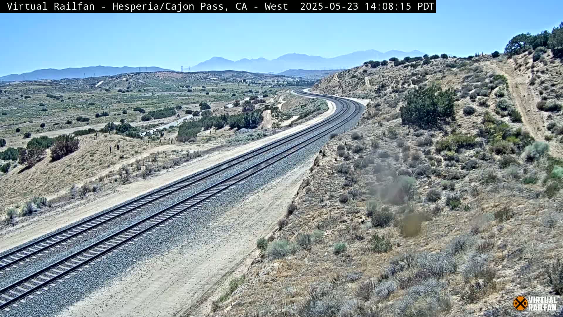 A long stretch of train tracks curves through a dry, desert-like landscape under a clear, sunny sky, with mountains visible in the distance.