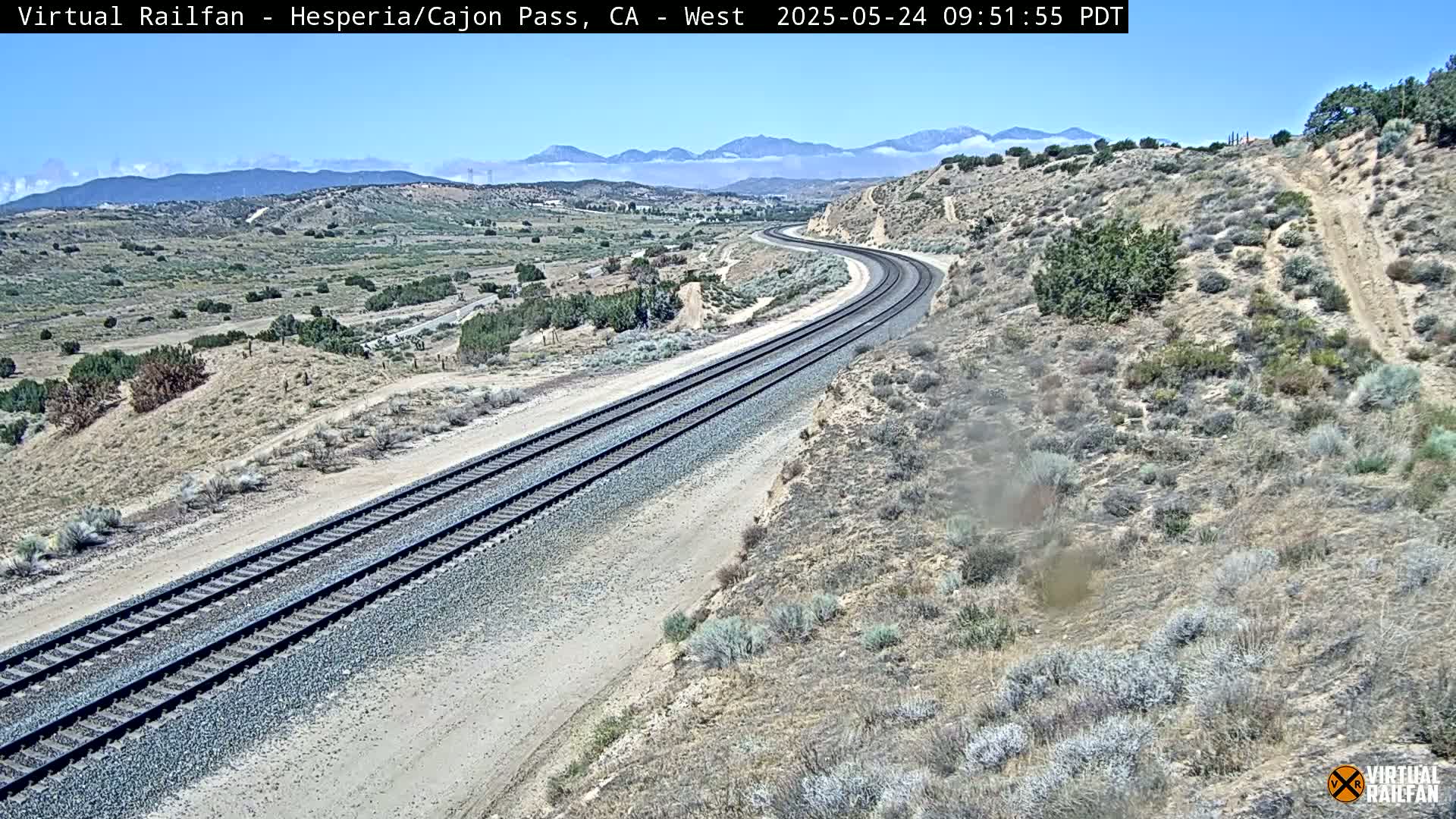 A long stretch of train tracks curves gently through a dry, desert-like landscape under a clear, sunny sky with distant mountains visible in the background.