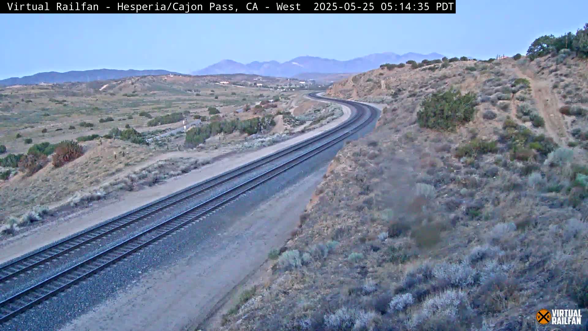 A high-angle view of a curving train track in a desert landscape under a clear, twilight sky.