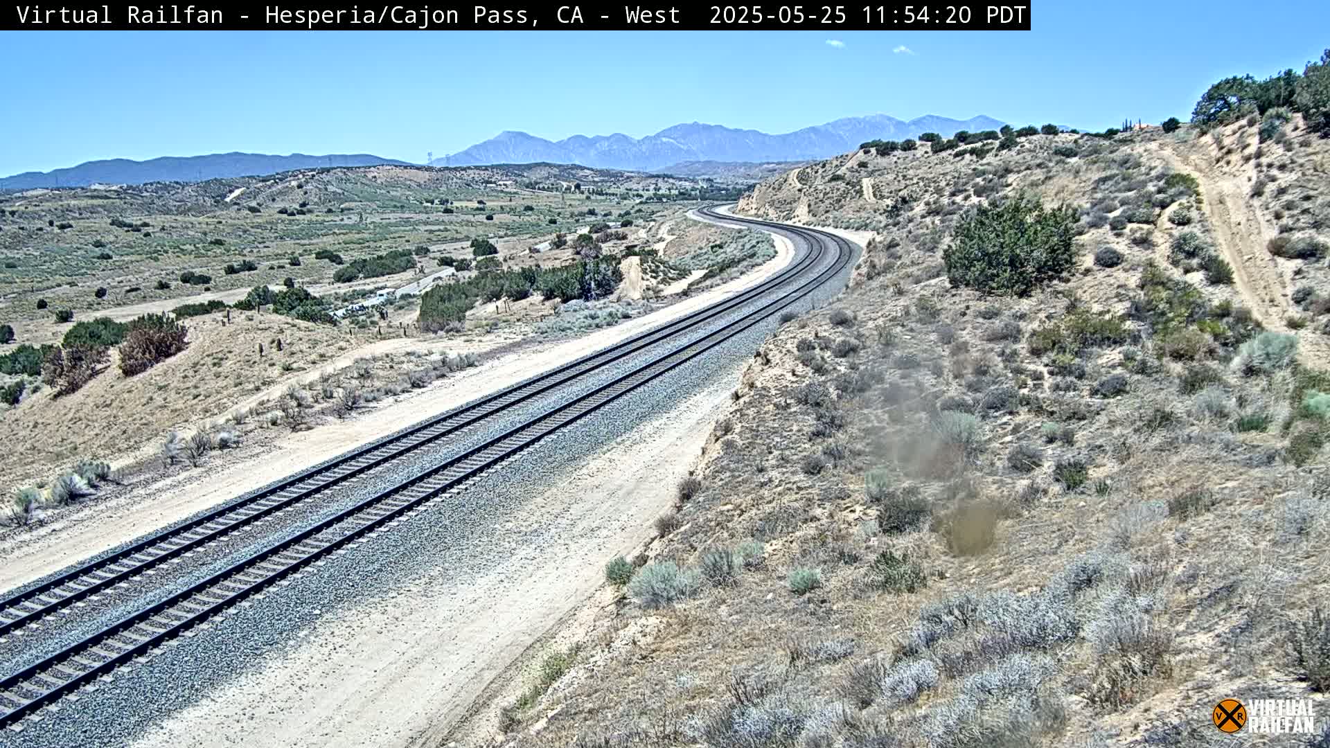 A long, curving railway track stretches across a desert landscape under a clear sky, with mountains visible in the distance.