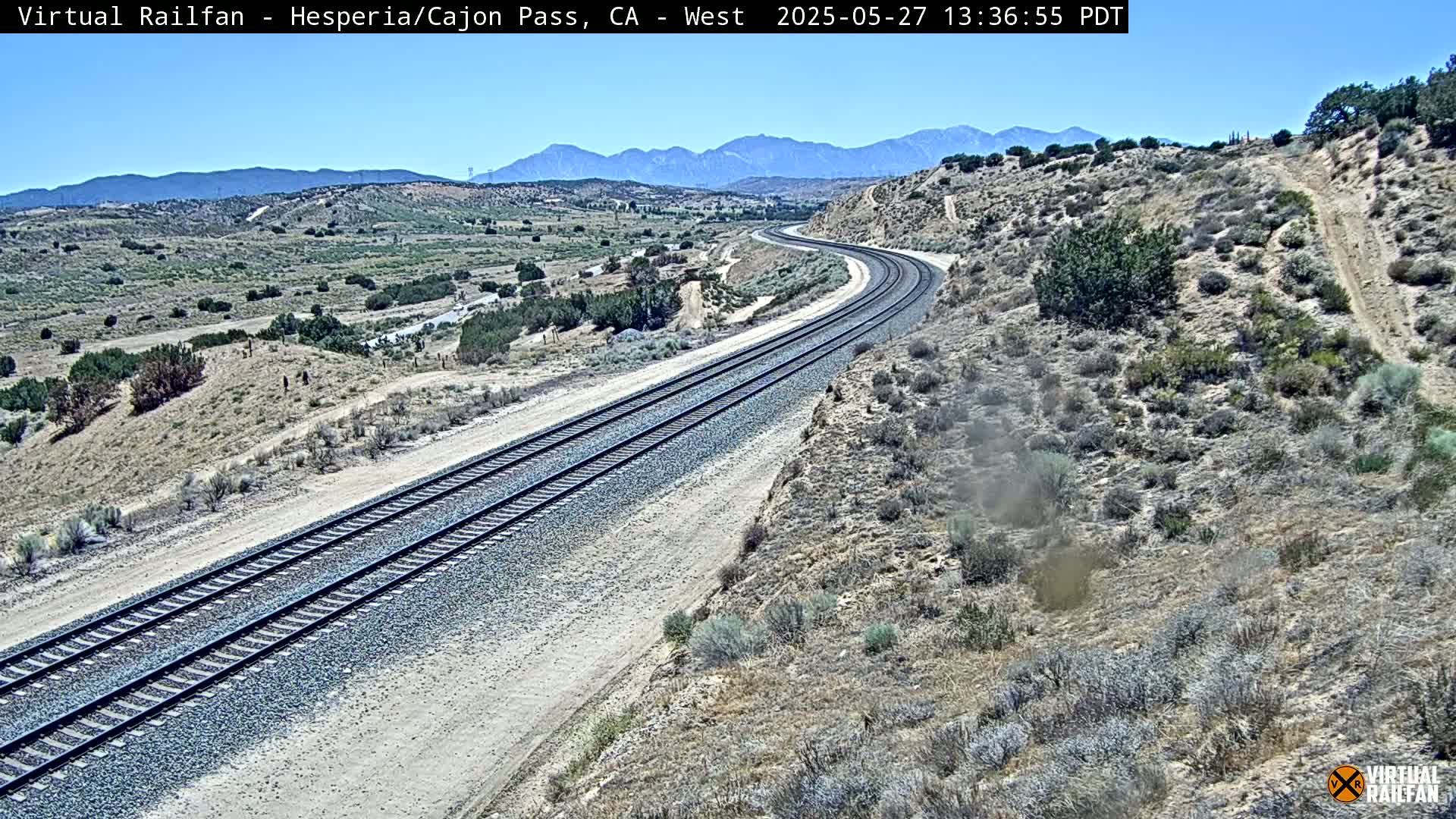 A long stretch of train tracks curves gently through a dry, desert landscape under a clear blue sky.