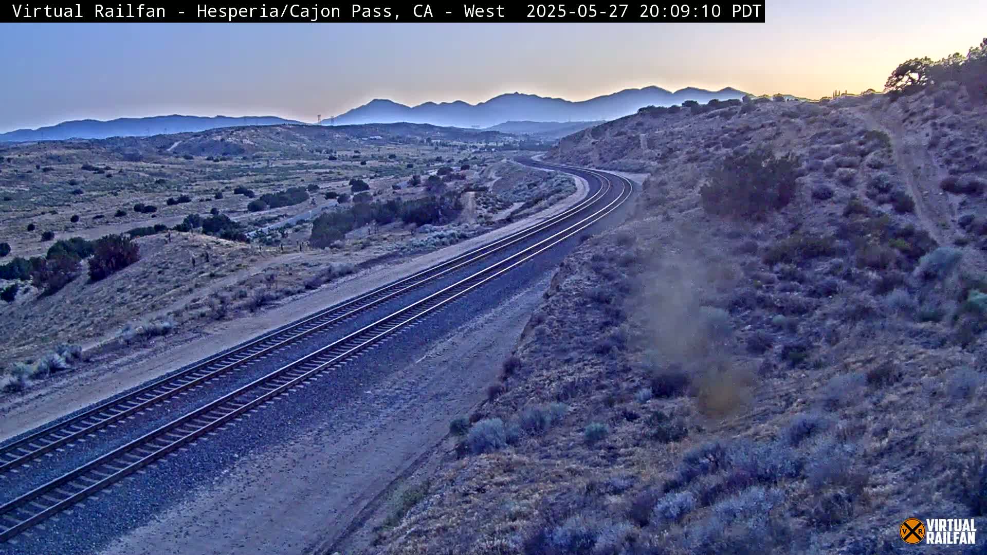 A high-angle, twilight view shows a pair of train tracks curving through a desert landscape under a clear sky.