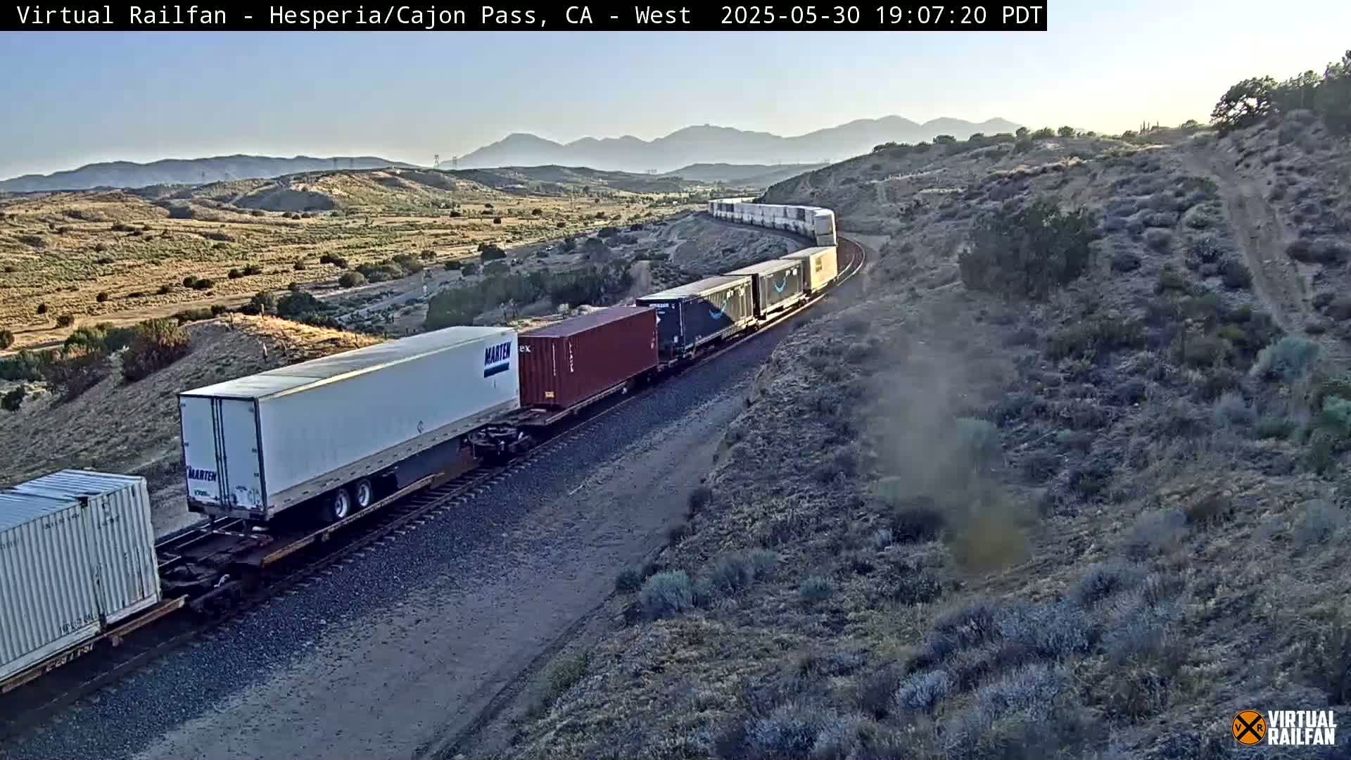 A long freight train carrying various shipping containers travels along a railroad track through a dry, mountainous landscape under a clear, twilight sky.