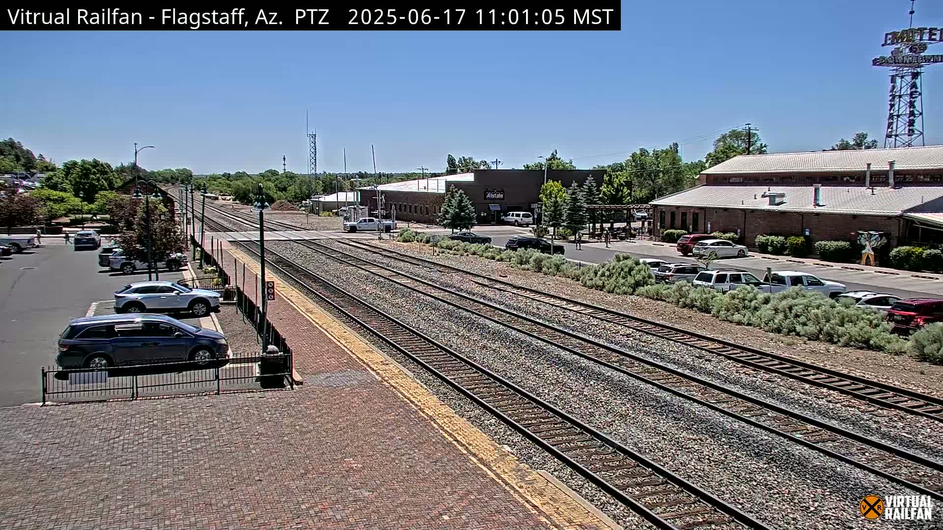 A sunny view of a train station with multiple sets of tracks, parked cars, and buildings in the background.