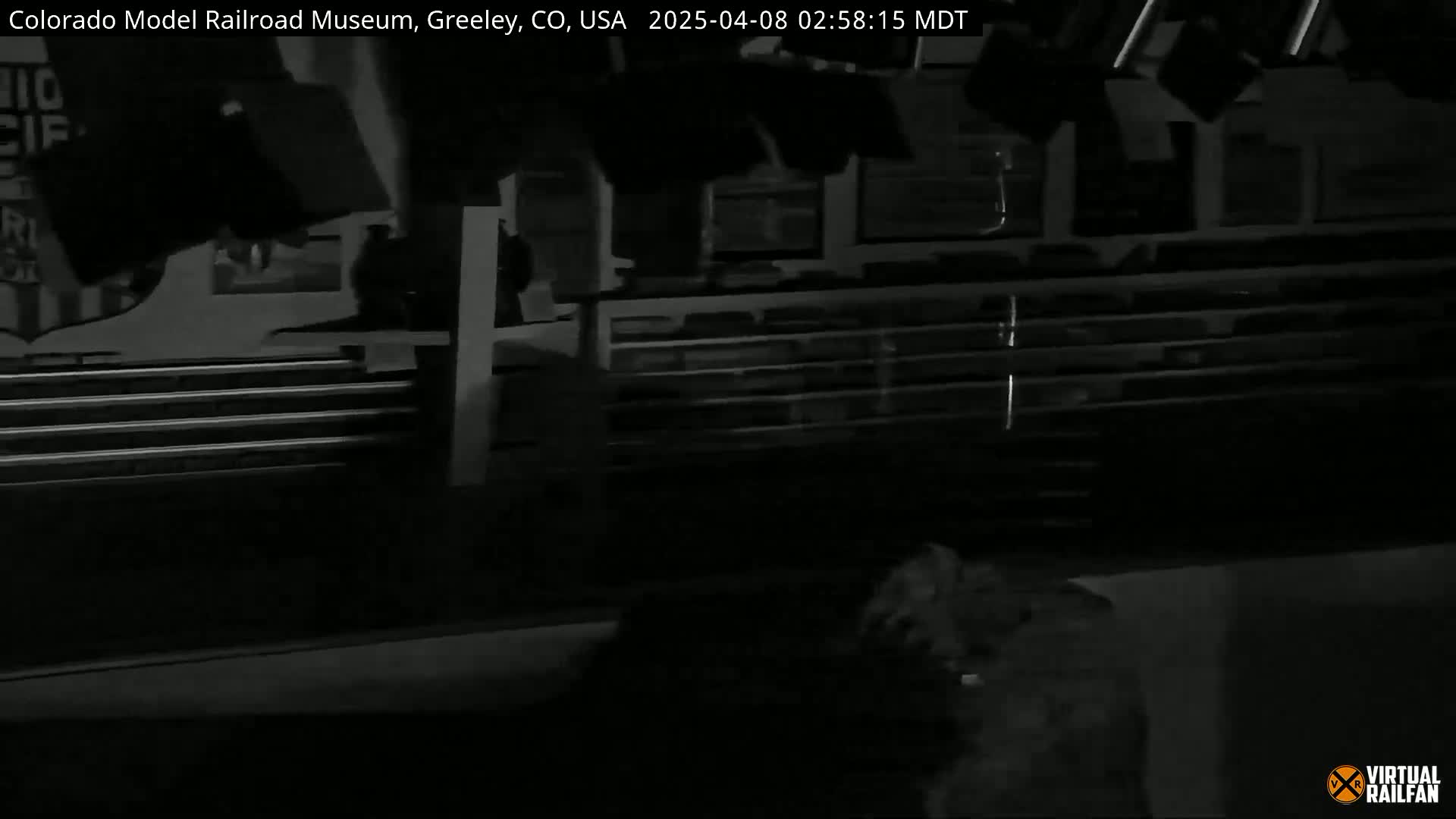 A dark, nighttime, grayscale image shows the interior of a building with dimly lit shelves and what appears to be a portion of a vehicle visible in the bottom left corner.
