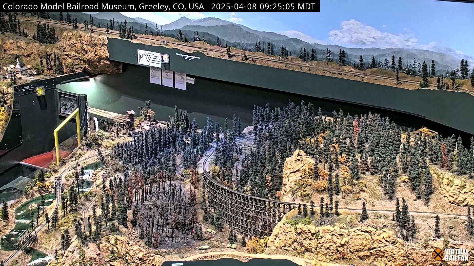 An extensive model railroad layout depicting a mountainous region with a winding track, a large trestle, and forested areas under a mostly clear sky.