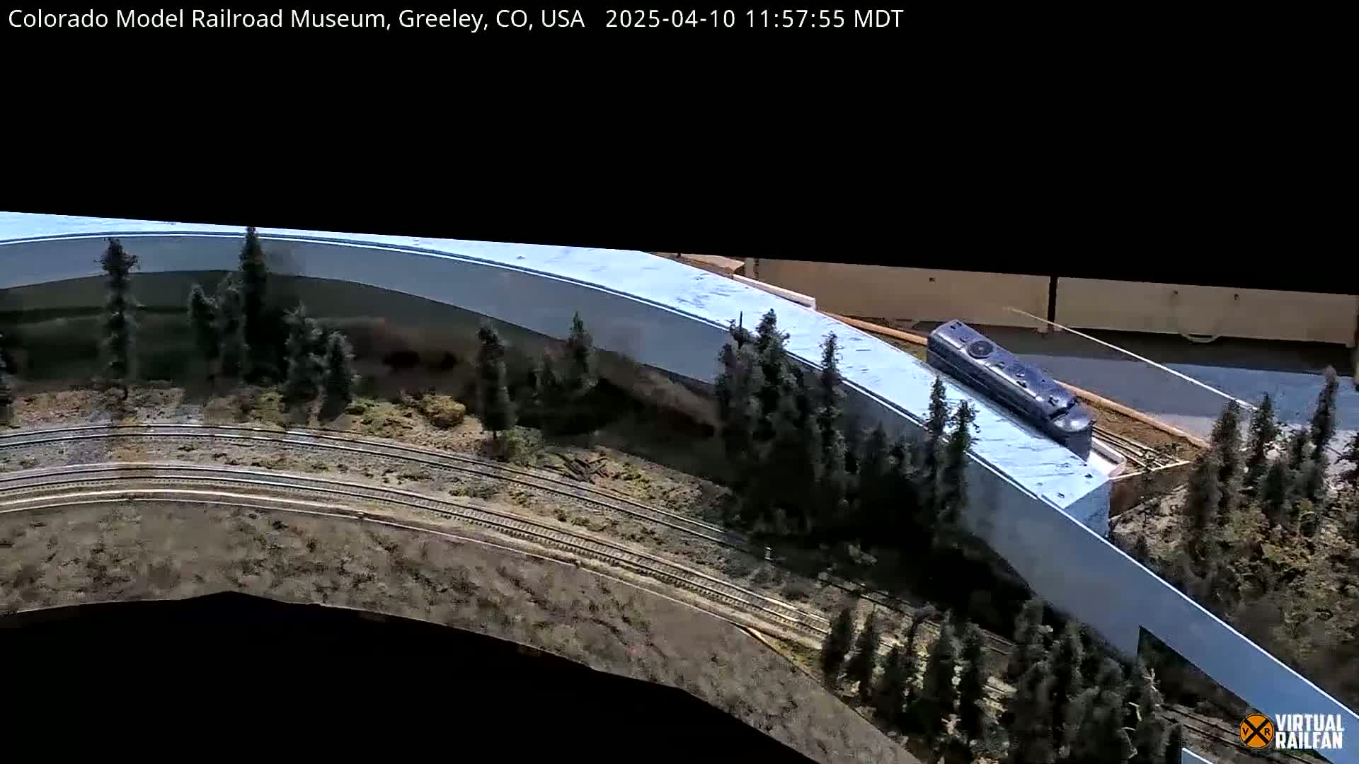 A model train curves around a model landscape featuring miniature evergreens and a light-blue structure.