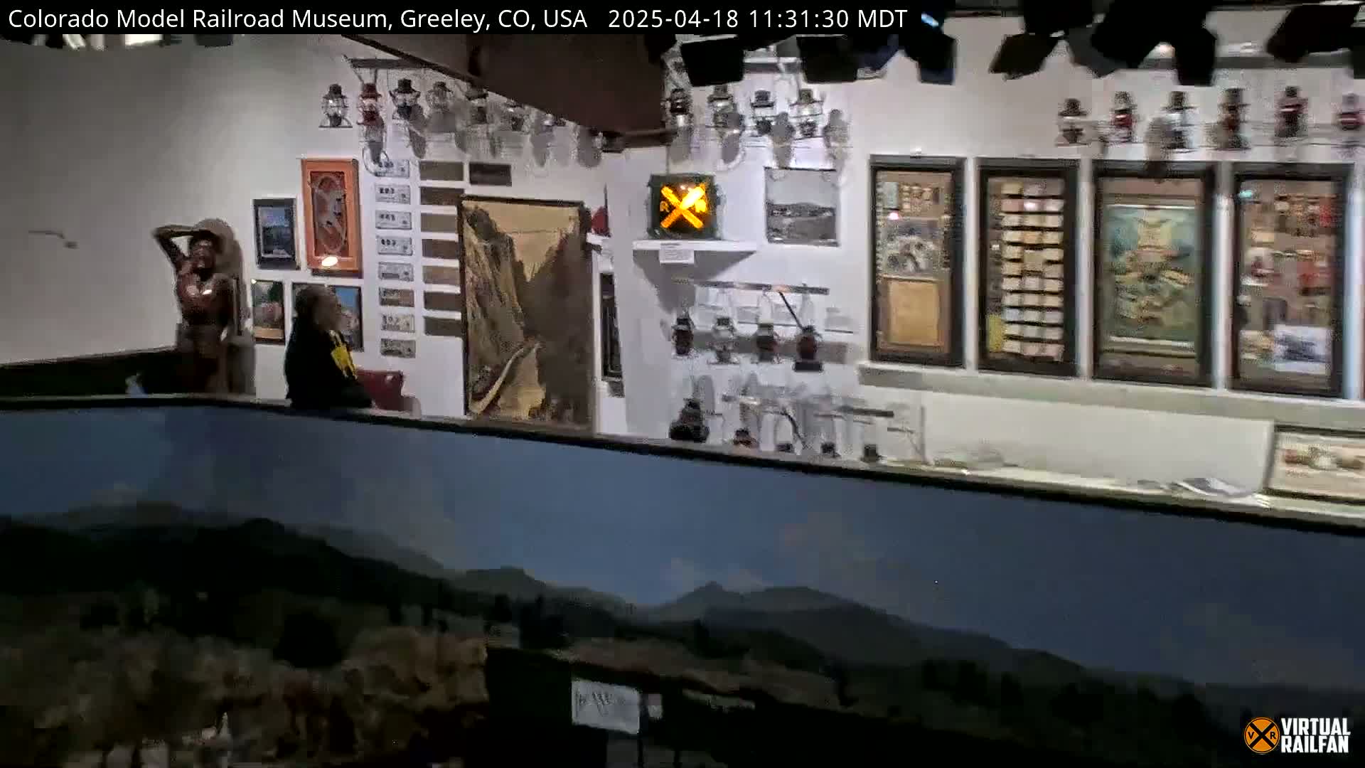 An indoor museum features various framed displays, including paintings and collections of artifacts, with a person standing near a wooden statue.