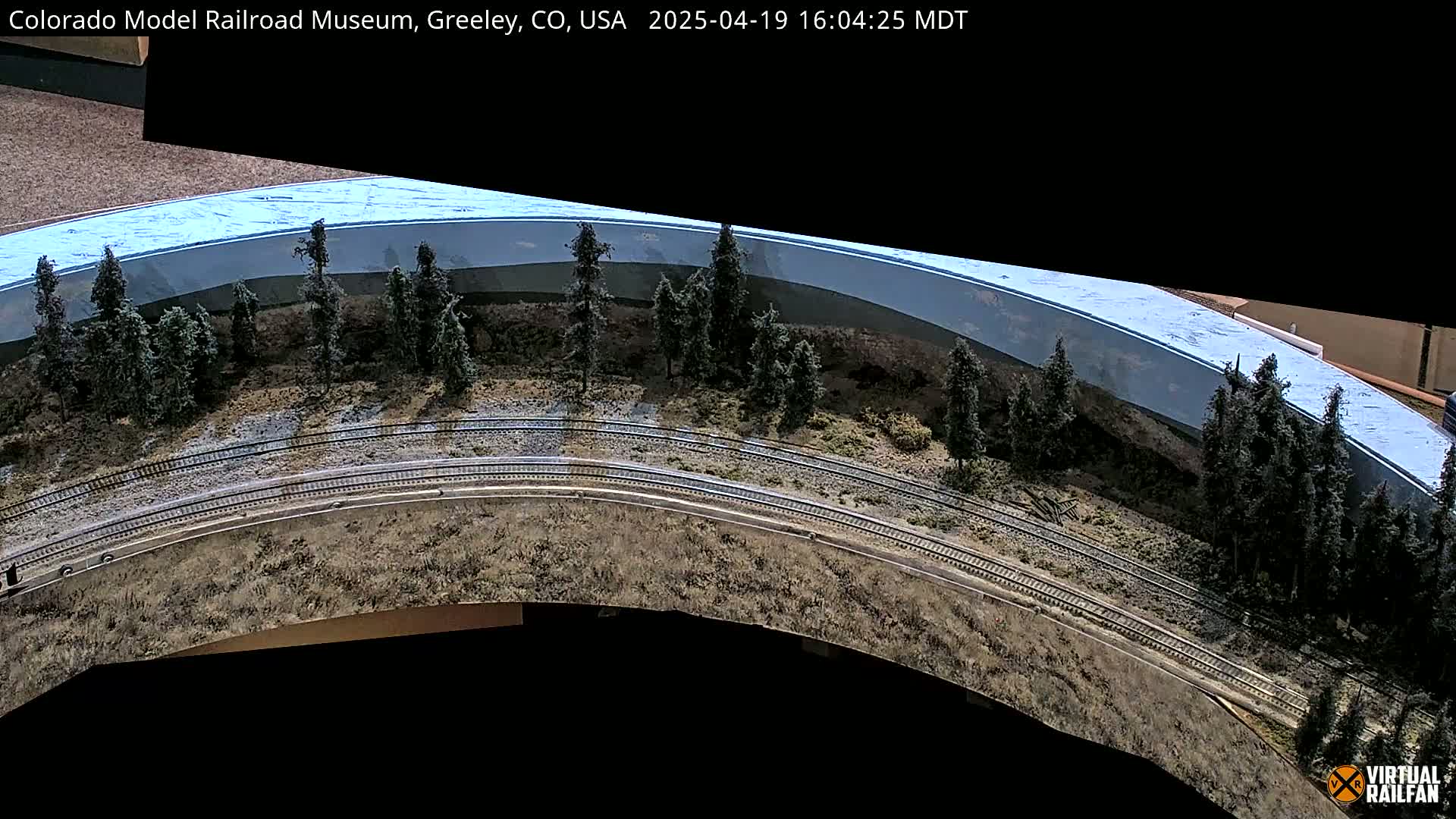 A model train track curves around a miniature landscape featuring evergreen trees and a light-blue painted backdrop.
