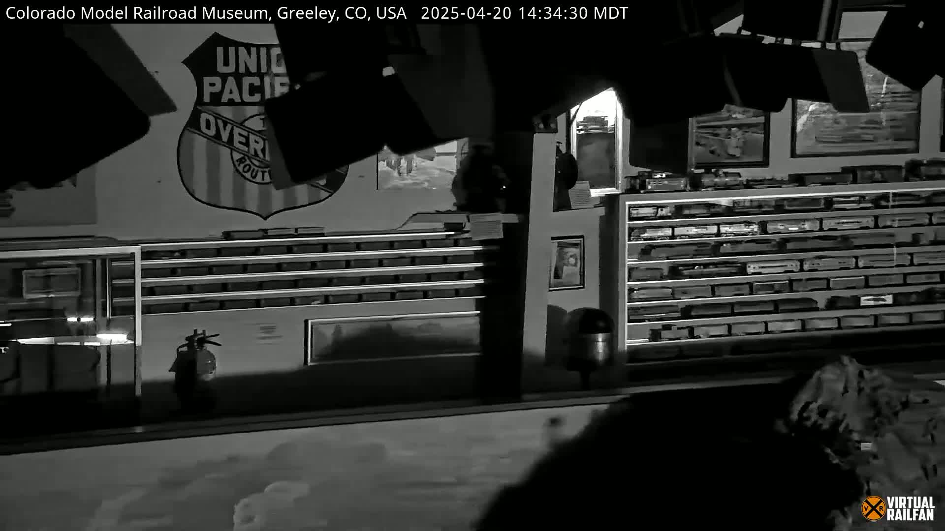 A dimly lit, black and white interior shows a large Union Pacific Railroad logo on the wall above numerous shelves filled with model trains.