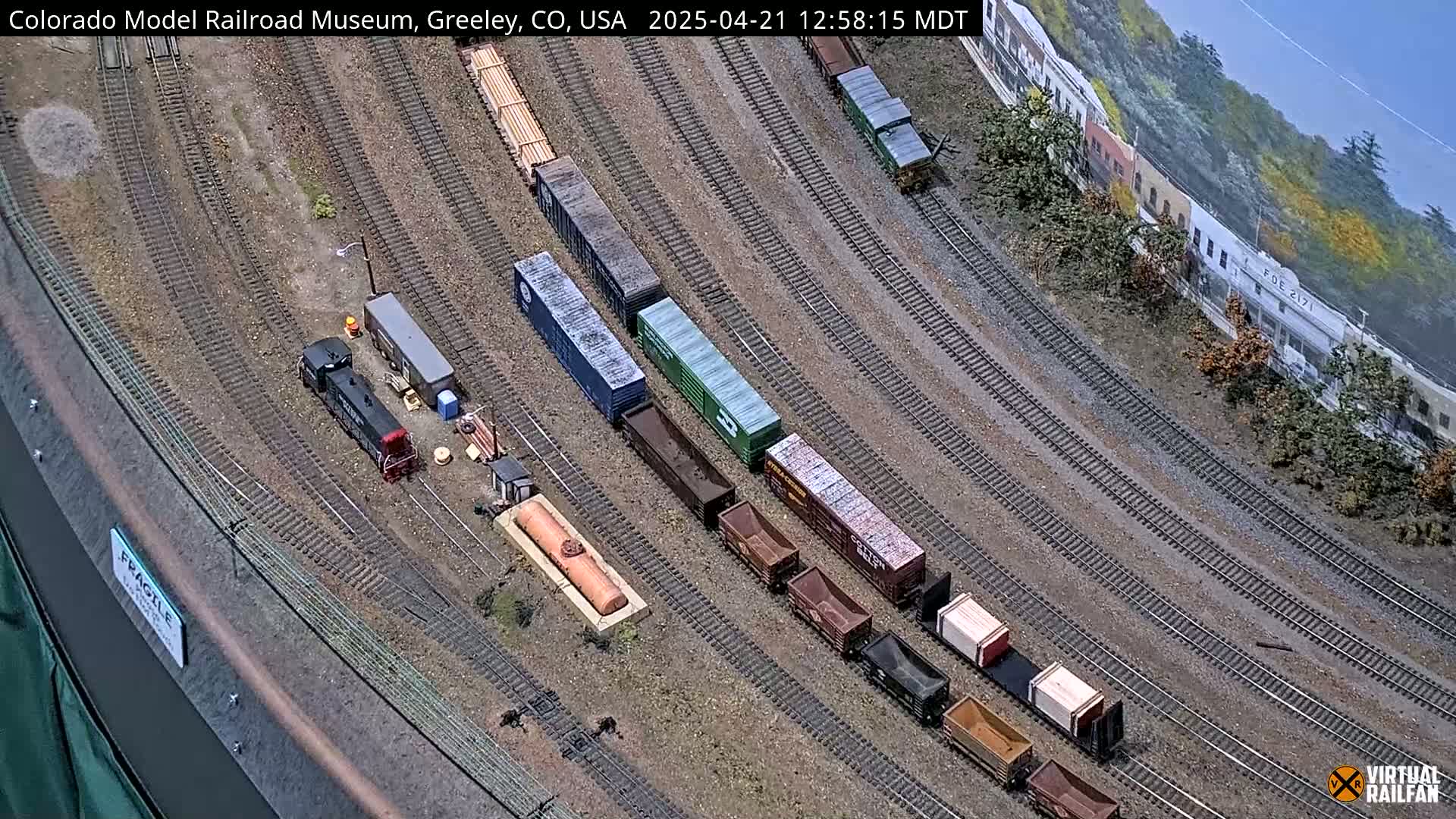 An overhead view of a model train layout shows multiple train cars and a locomotive on various tracks, set against a backdrop of miniature buildings and trees under a sunny sky.