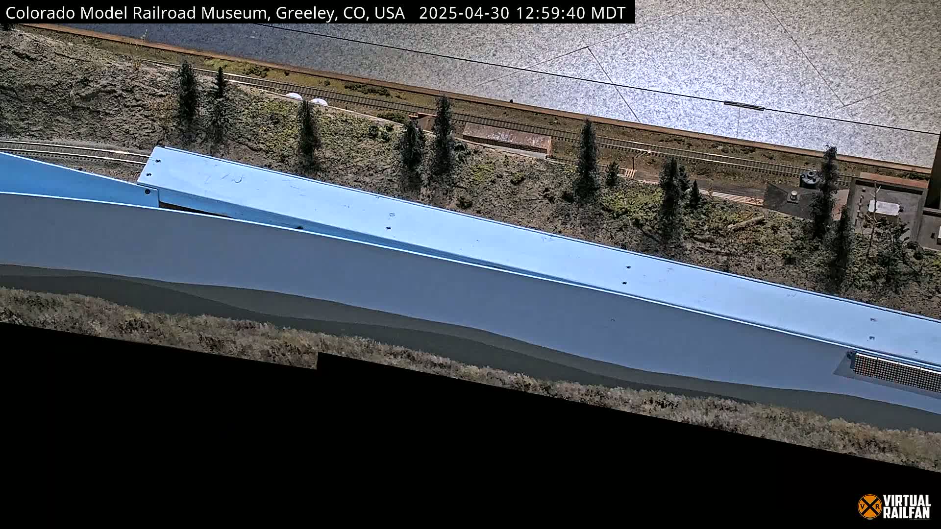 A high-angle view of a model railroad layout depicting a light-blue structure next to train tracks and miniature evergreen trees, near a body of water under sunny conditions.