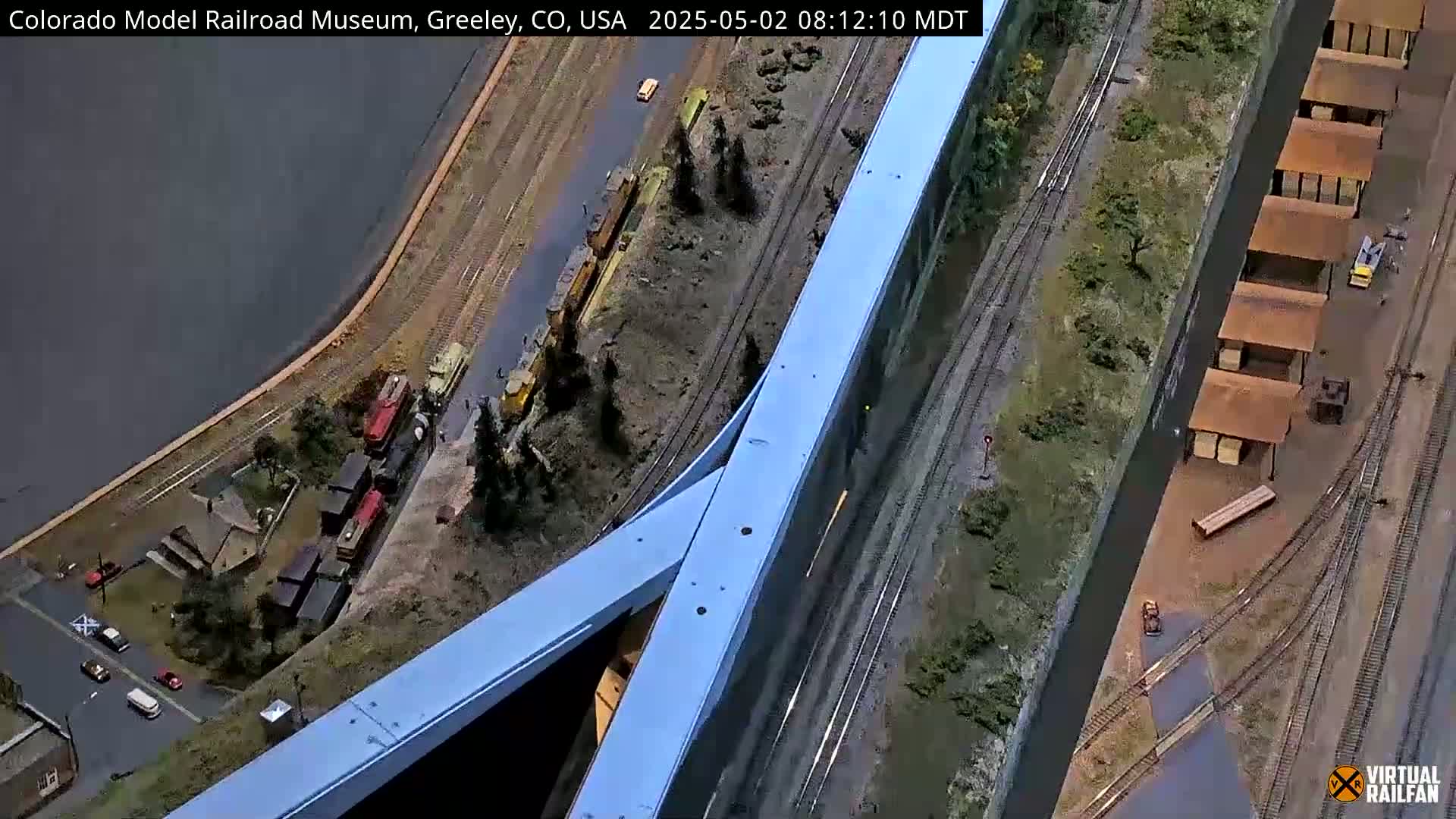 A detailed model railroad layout features multiple train cars, buildings, and tracks alongside a body of water.
