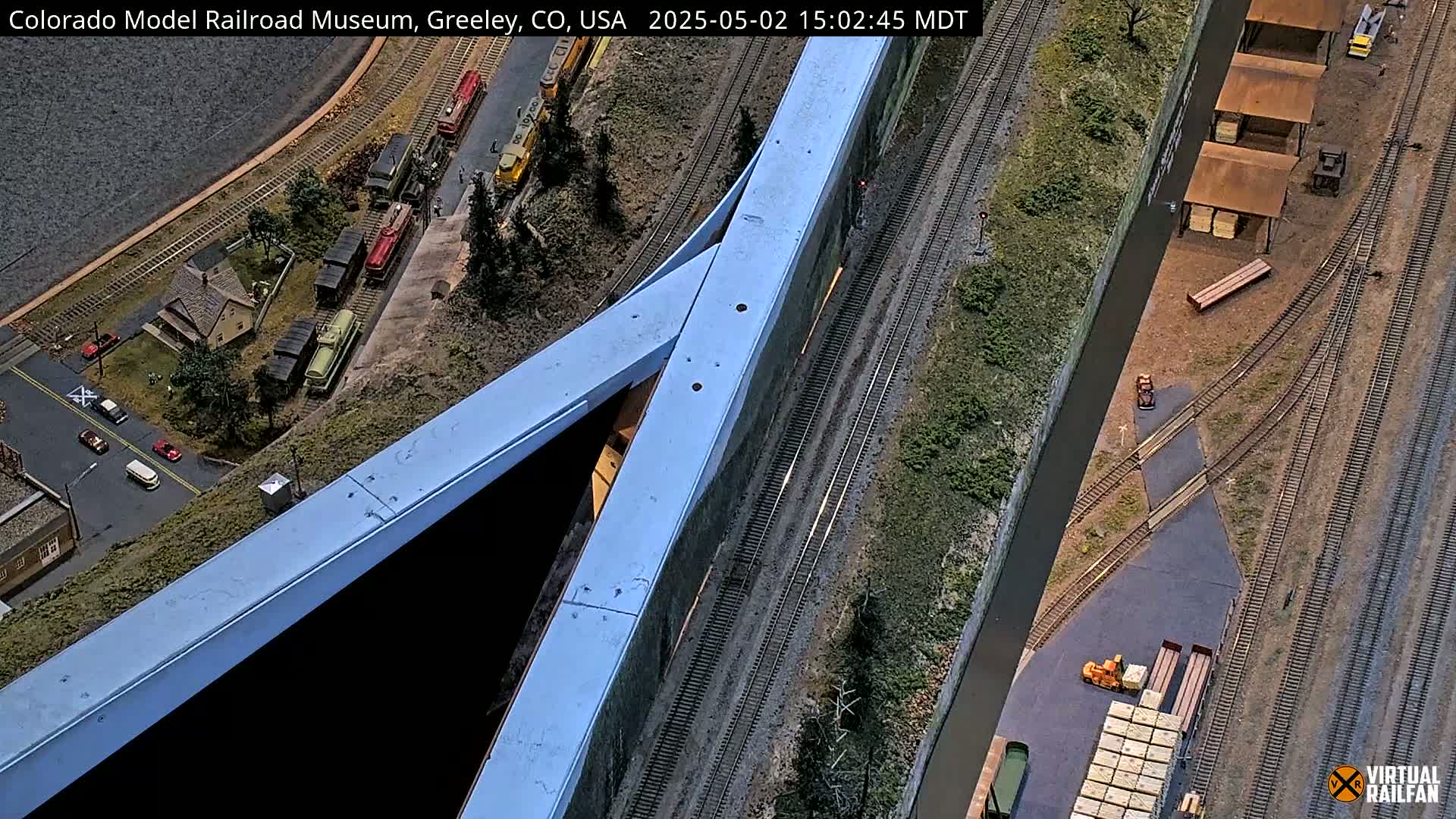 An overhead view of a detailed model railroad scene with trains, buildings, roads, and a large blue structure.