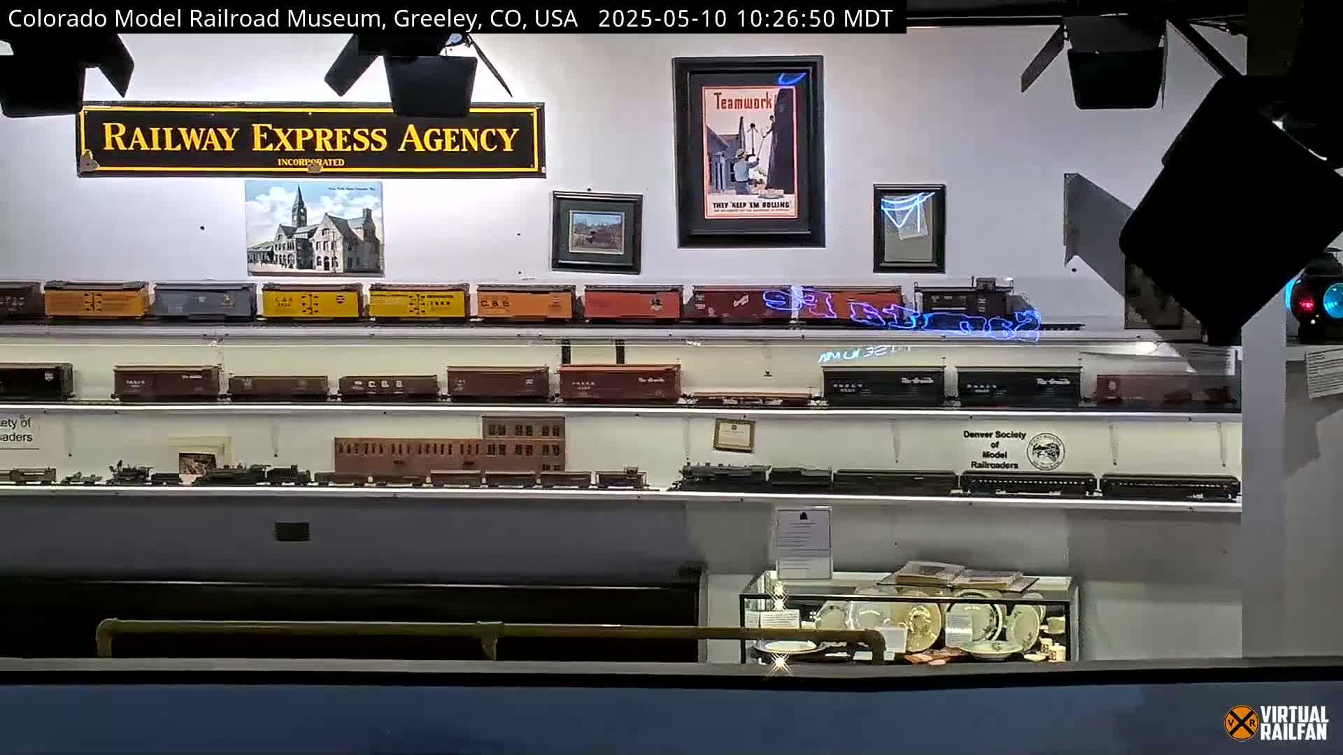 An indoor museum displays numerous model trains on shelves, along with framed posters and pictures.
