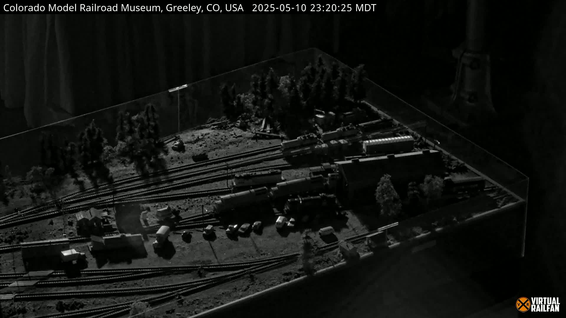 A detailed model railroad display featuring multiple trains, tracks, and miniature buildings, set within a glass enclosure.