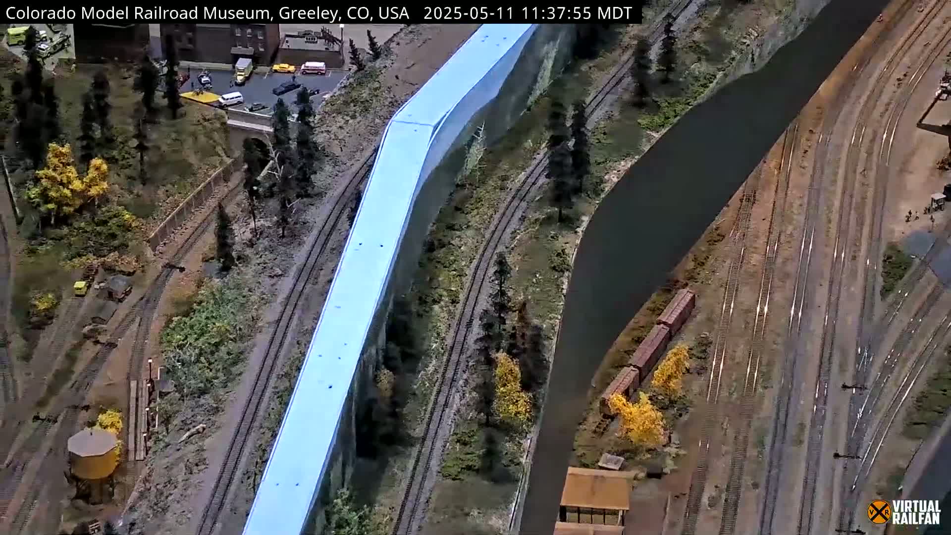 An overhead view of an extensive model railroad layout with multiple train tracks, a large light-blue structure, and miniature landscaping, including trees and buildings.