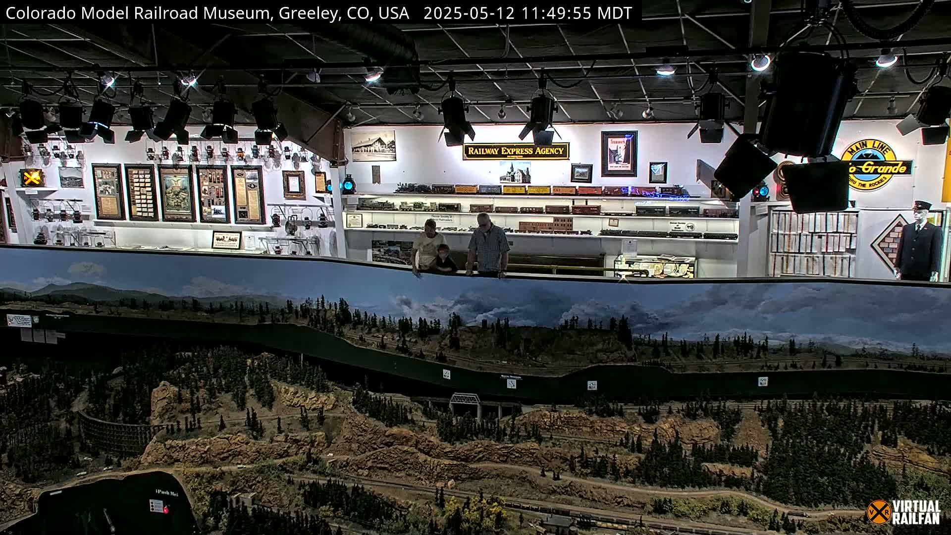 An indoor model railroad display featuring a large, detailed landscape backdrop with mountains and forests, observed by three people.
