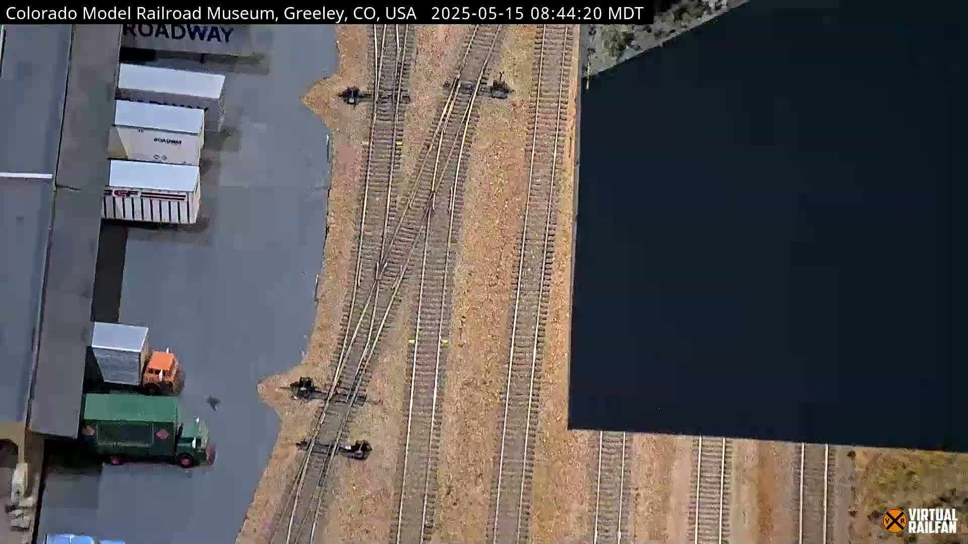 An aerial view shows a model railroad switching yard with several boxcars and trucks parked nearby.