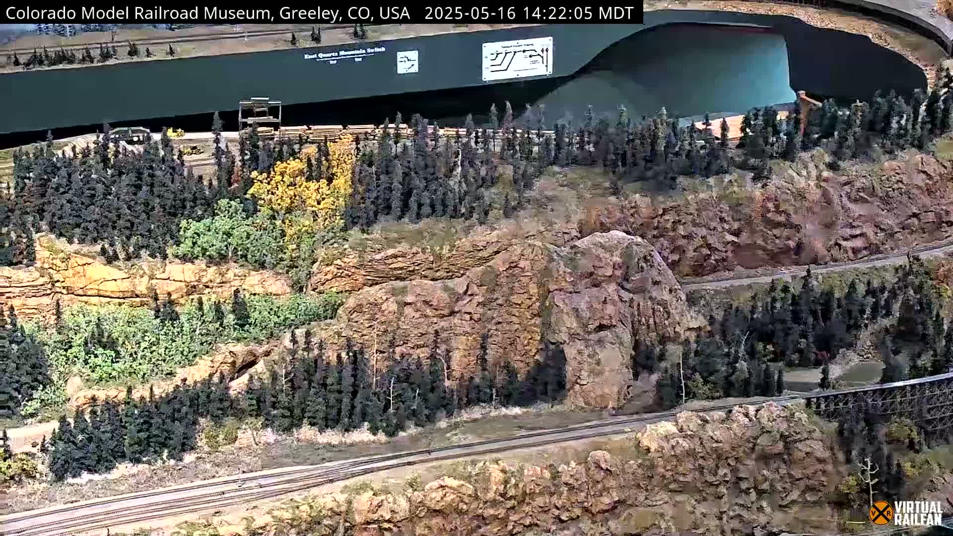 A large model railroad display features a winding track running through a miniature landscape of rocky cliffs, forests, and a body of water.