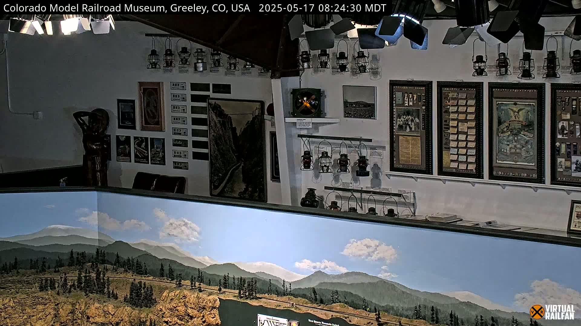 An indoor museum display features numerous antique lanterns, framed photographs and documents, a statue, and a large painted backdrop of a mountain landscape under a partly cloudy sky.