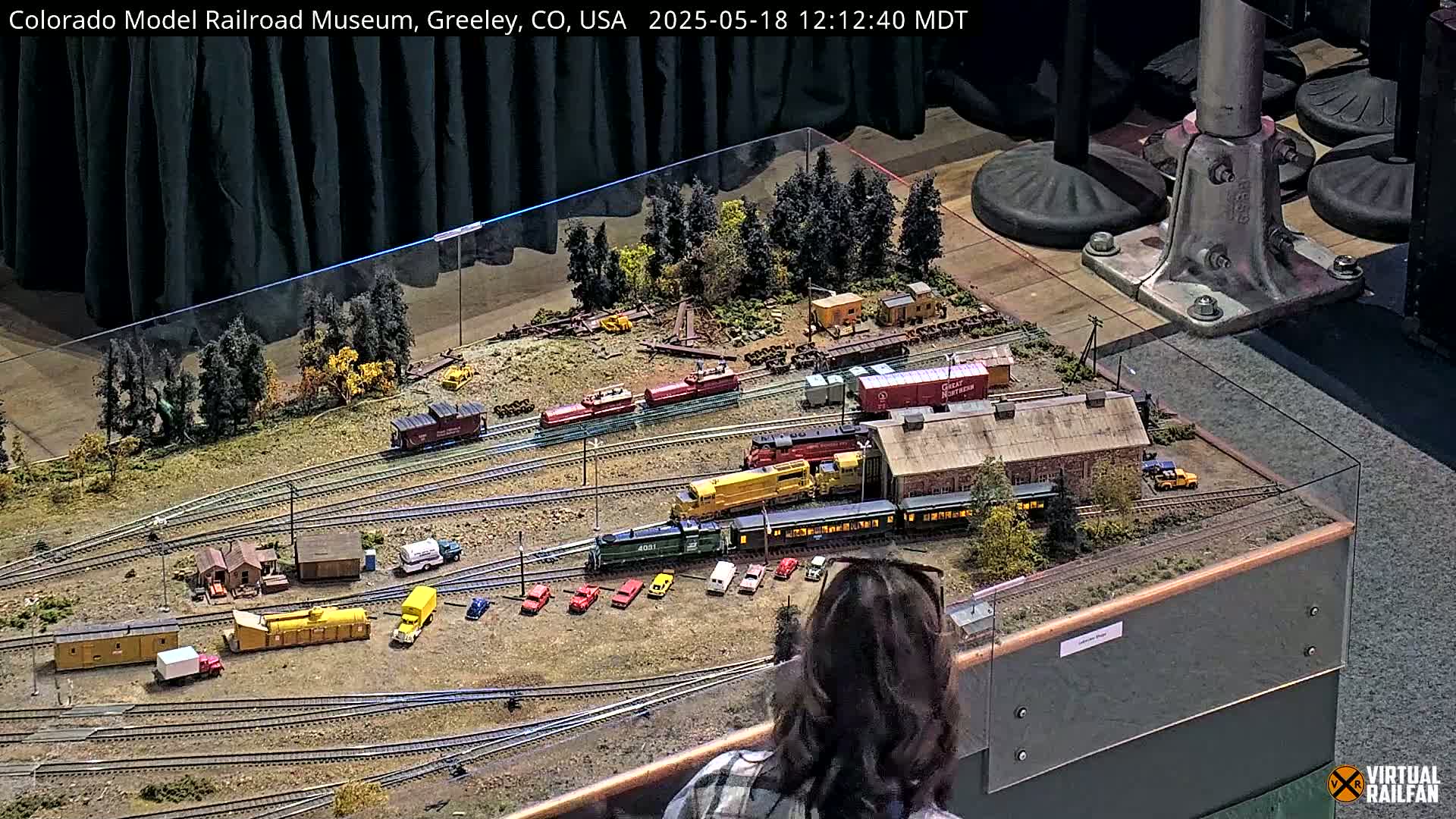 A large, detailed model railroad display with multiple train cars, buildings, and vehicles is viewed from above by a person with brown hair.
