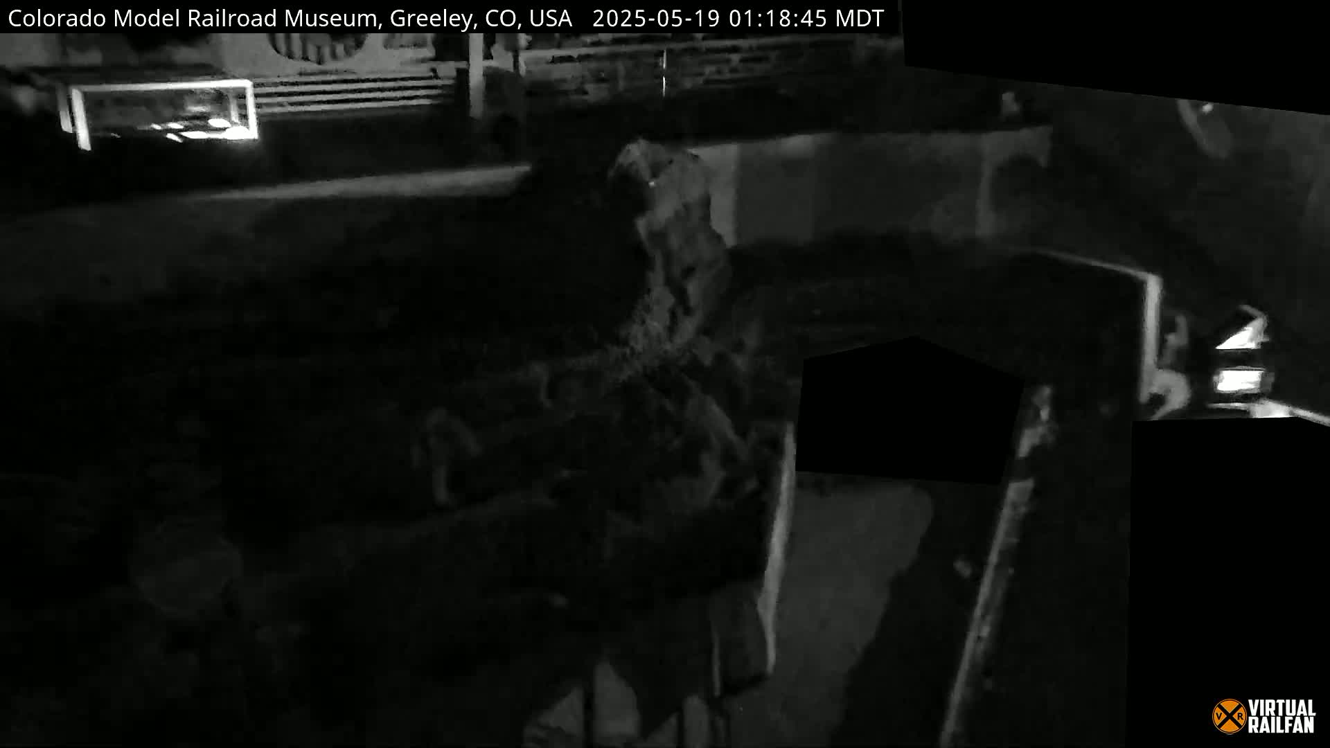 A dimly lit, dark grayscale image shows an indistinct object, possibly a piece of machinery, within a shadowed interior space.