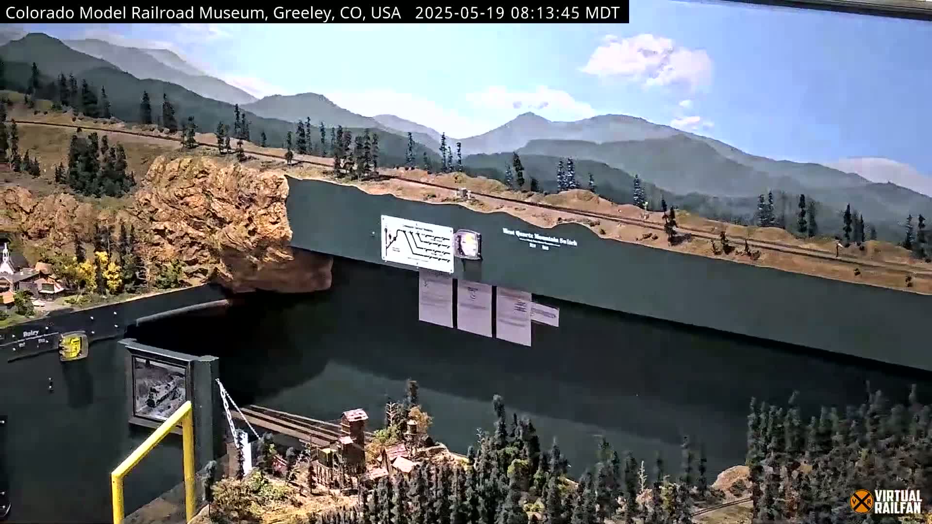 A large model railroad scene depicting a mountain landscape under a partly cloudy sky.