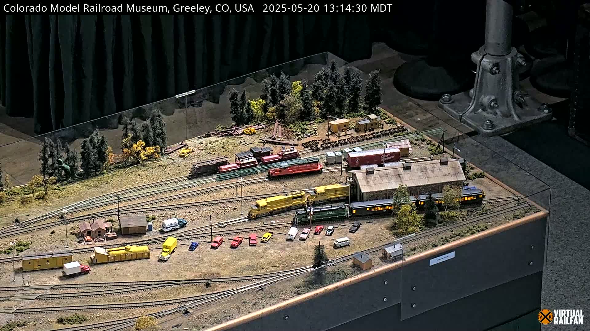 An intricate model railroad display features multiple train cars, buildings, and vehicles arranged on a detailed landscape with miniature trees.