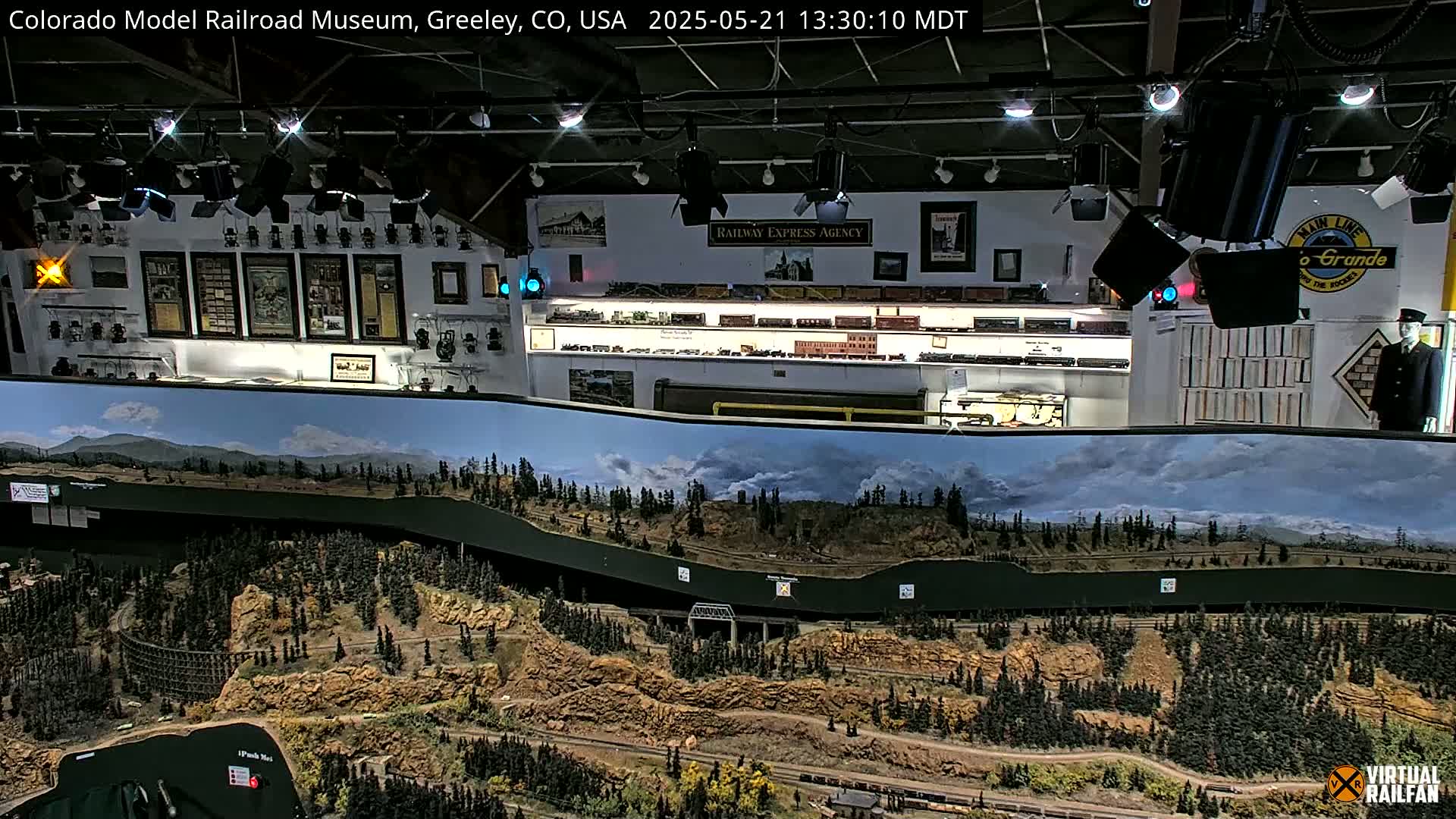 An indoor model railroad display features a large, detailed landscape diorama of a mountainous region with a train track winding through it, and a wall filled with railroad memorabilia.