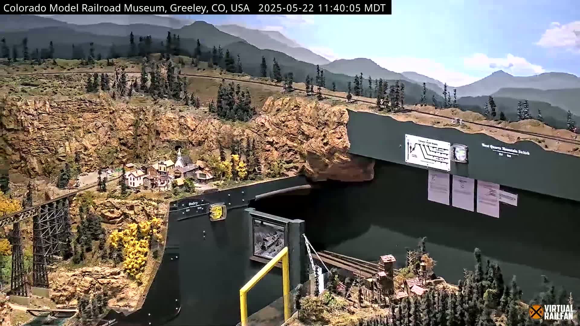 An expansive model railroad layout depicting a mountainous region with a town, a trestle, and tunnels, all under a clear, sunny sky.