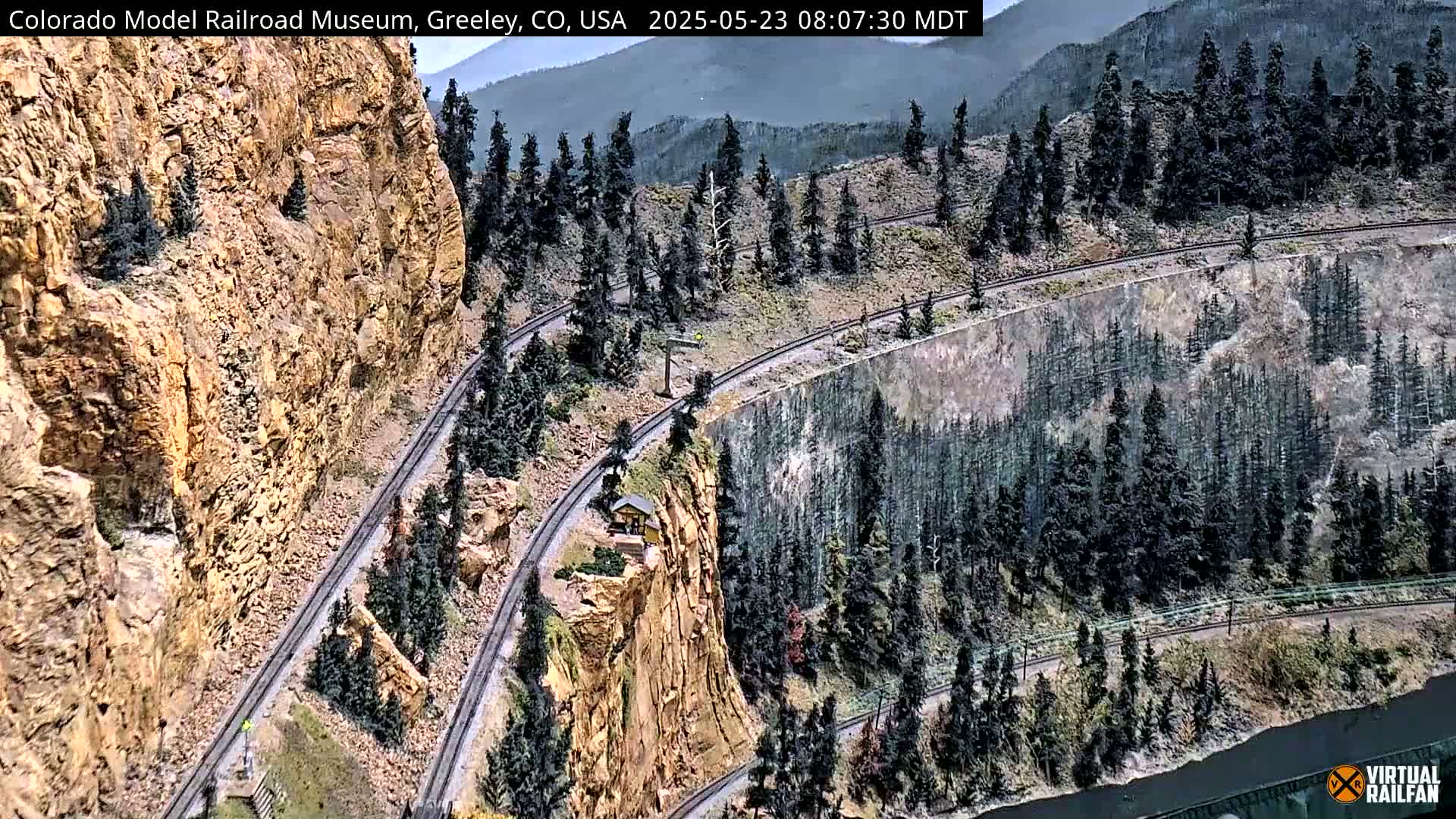 A detailed model train layout depicting a mountainous landscape with winding tracks, a small building, and evergreen trees under a partly cloudy sky.