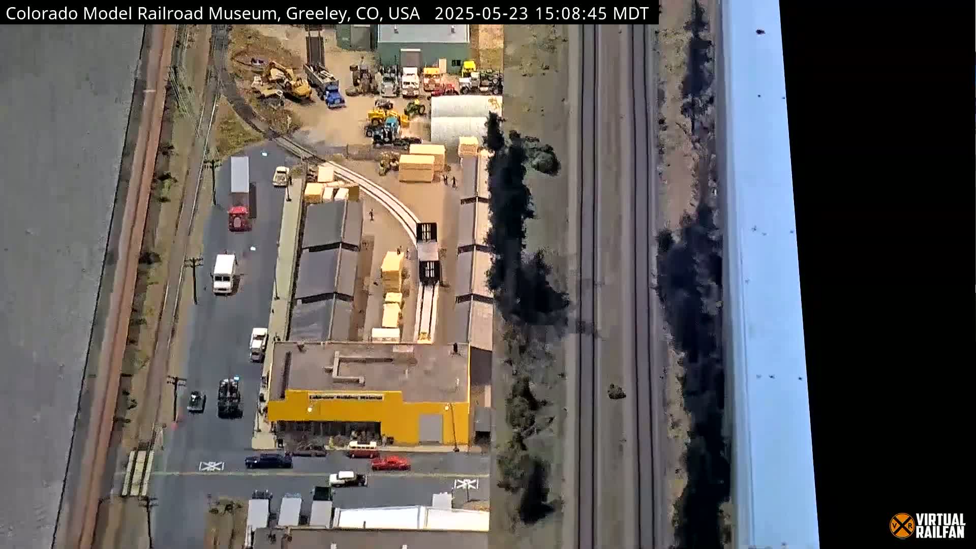 An aerial view shows a model railroad museum, surrounding buildings, vehicles on a road and nearby train tracks on a sunny day.
