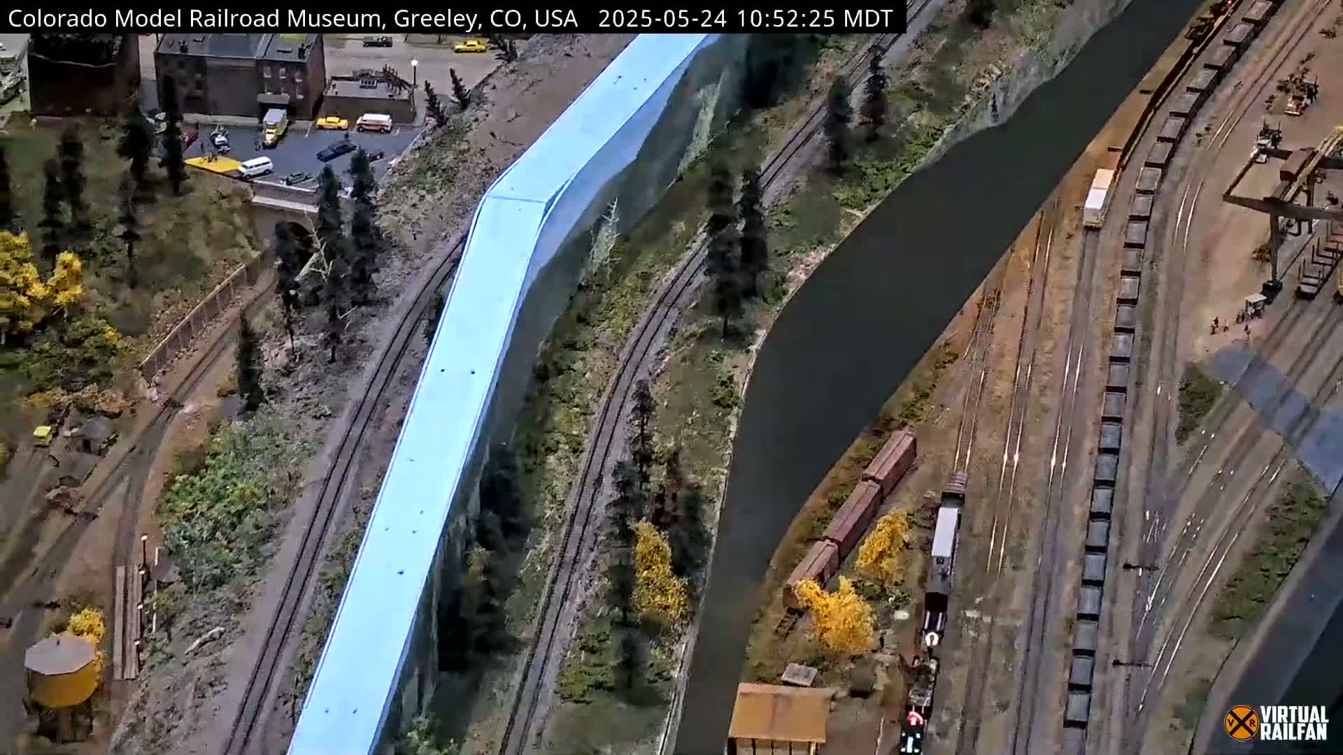 An overhead view of a model railroad with several trains, tracks, and miniature buildings, situated beside a simulated mountainside.