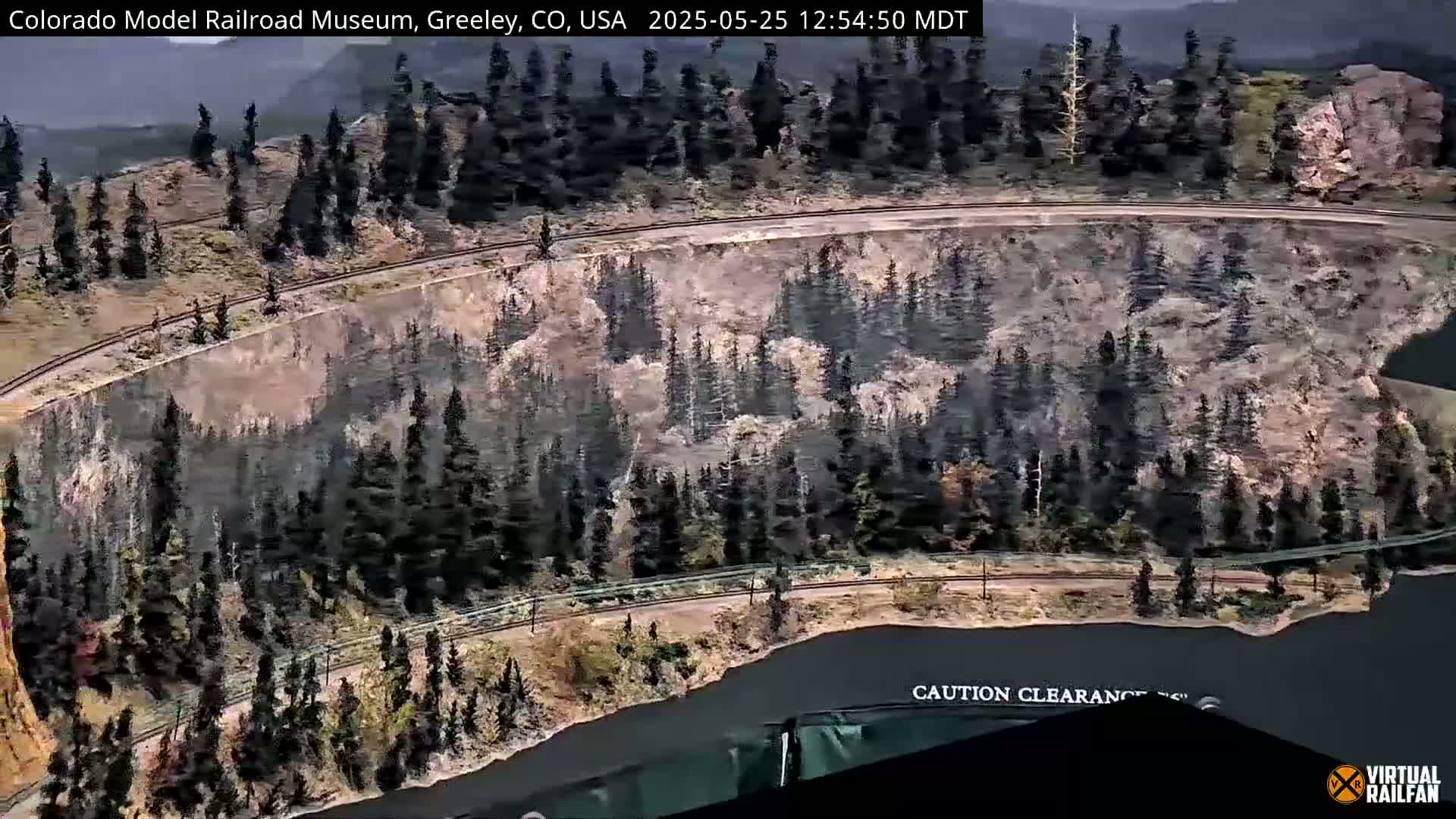 An overhead view shows a model train track curving around a hillside covered in evergreen trees under a partly cloudy sky.