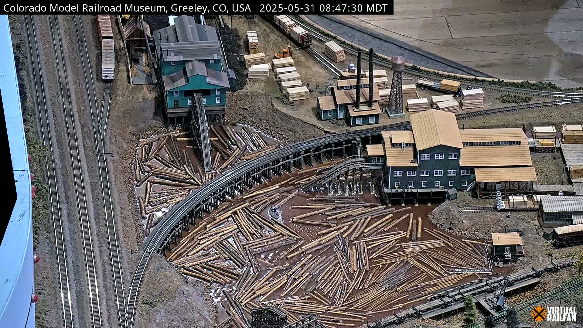 An overhead view of a model train layout depicting a lumber mill with logs floating in water, alongside railway tracks and buildings.
