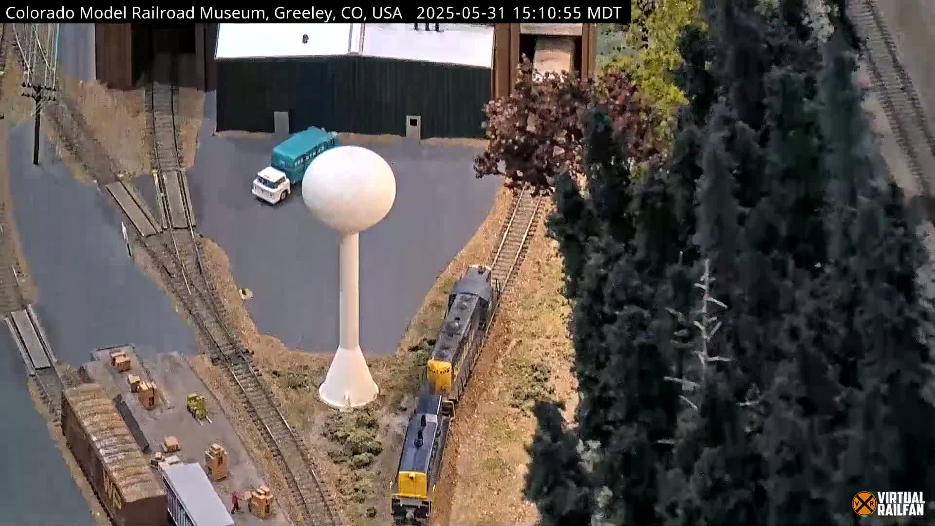 A model train travels alongside a water tower and several trucks near a loading dock, next to evergreen trees.