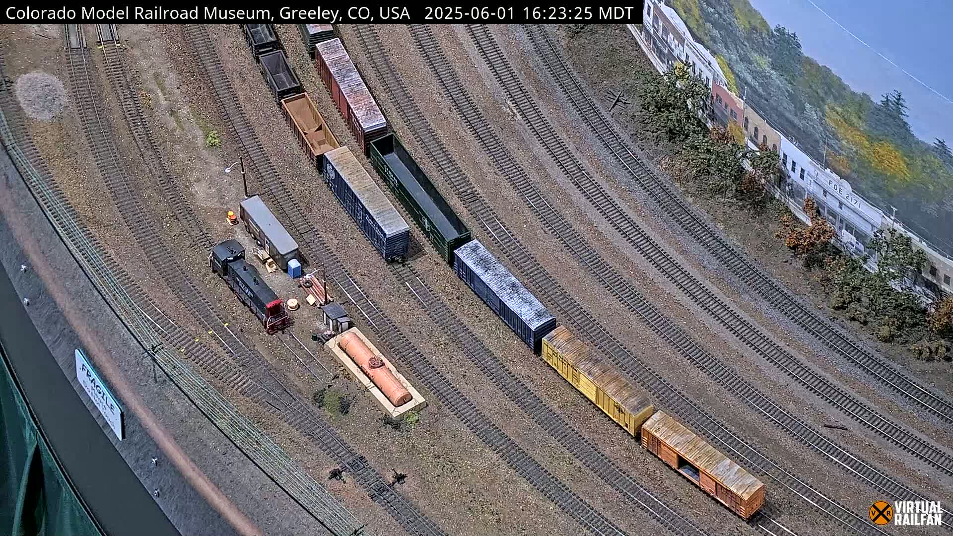 An overhead view of a model train set displays numerous rail cars and a locomotive on a curving track, set against a backdrop of miniature buildings and foliage under a clear sky.