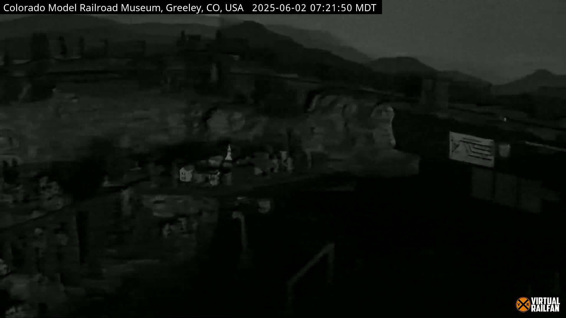 A dark, nighttime view of a model railroad display, showing a miniature town and landscape under a mostly cloudy sky.