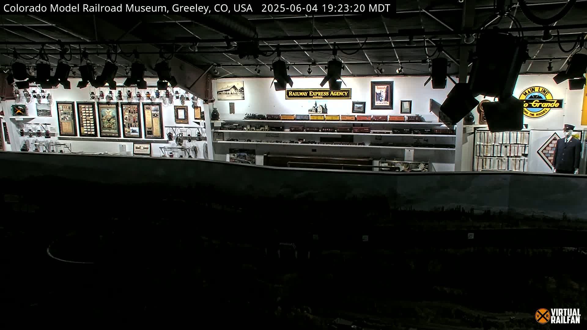 A dimly lit indoor model railroad museum displays various model trains, framed artwork, and railroad memorabilia on shelves and walls.