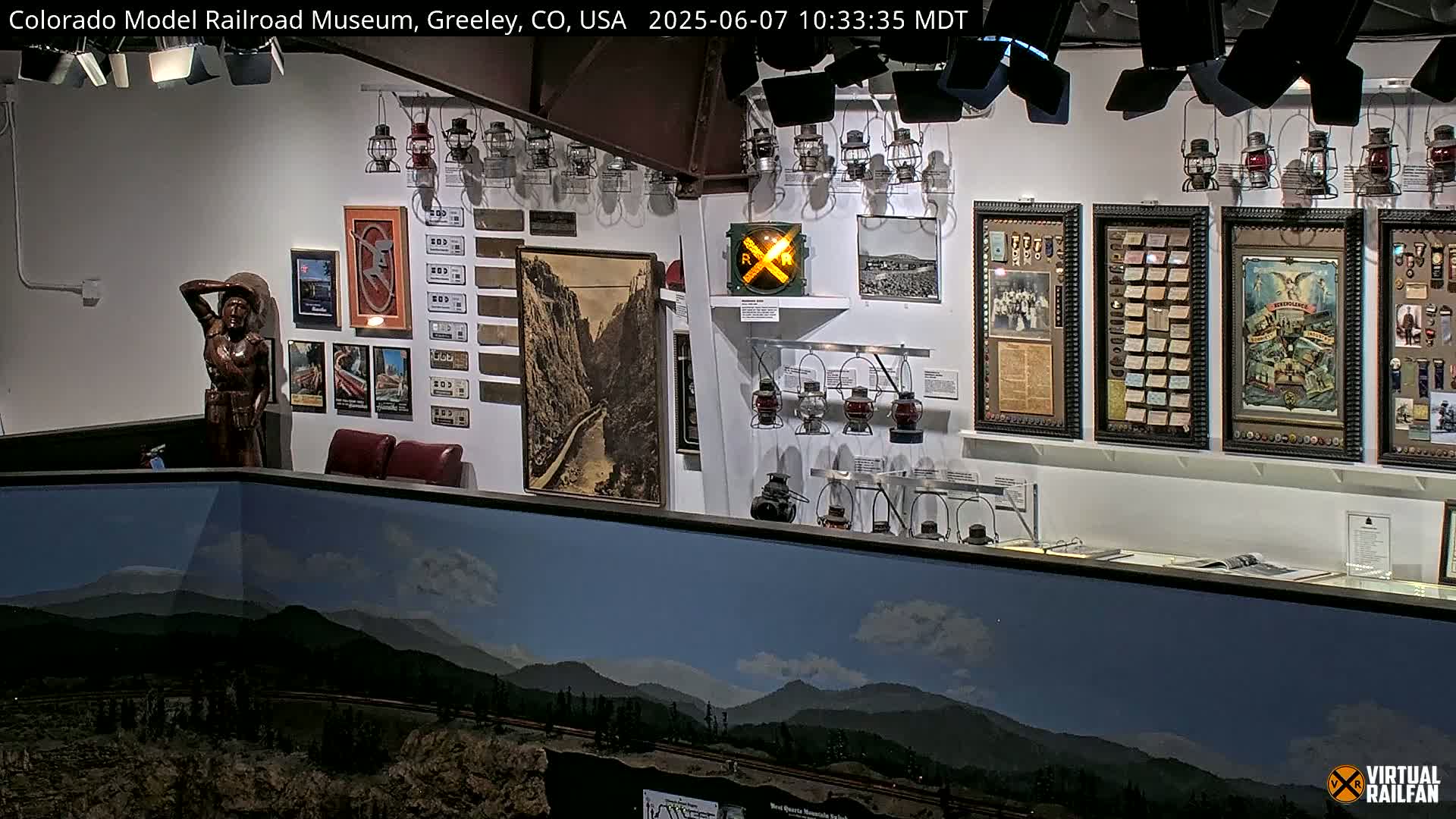 A museum interior displaying various railroad artifacts, including lanterns, photographs, and a wooden statue, with a large landscape painting of mountains visible in the foreground.