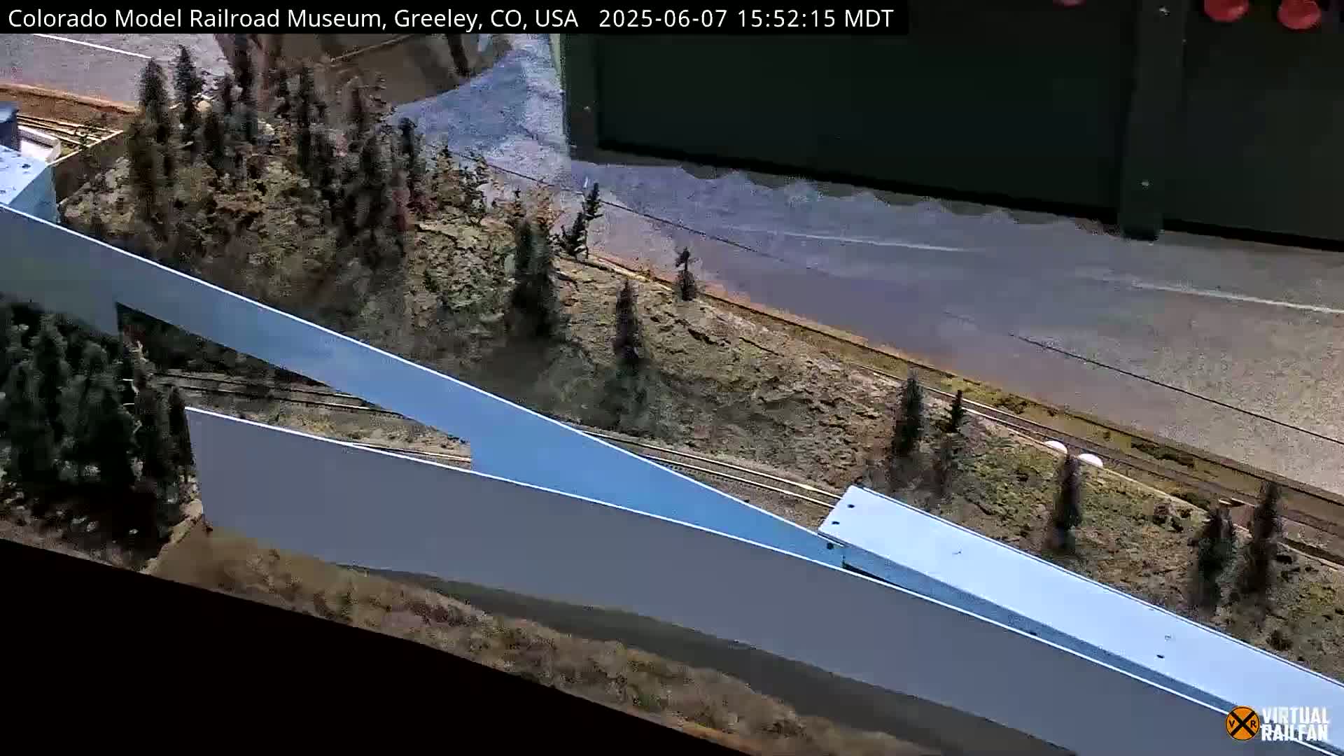 A model train travels along tracks beside a light-blue wall, passing a miniature landscape of trees and rock formations.