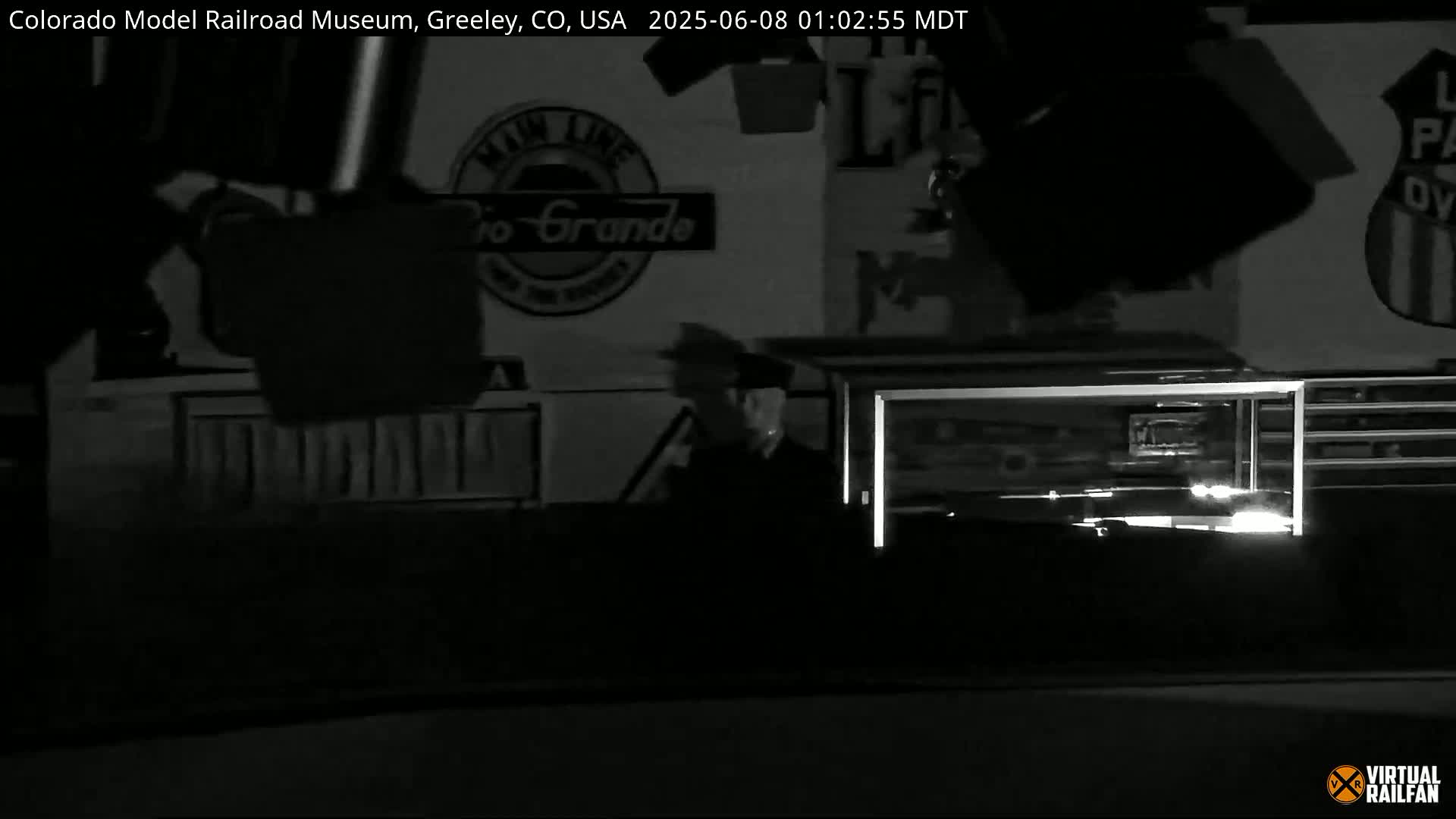A nighttime, black-and-white security camera image shows a person standing near a wall with Rio Grande signage and other indistinct objects.