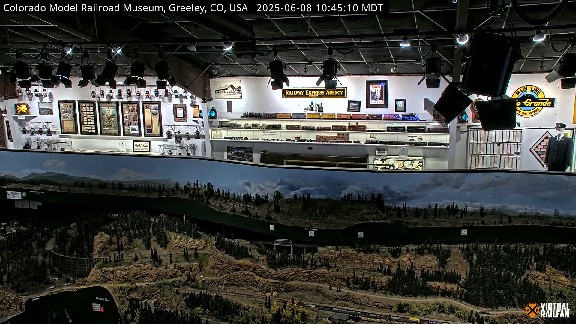 An indoor model railroad display features a large, detailed landscape diorama of a mountainous region with a train track winding through it, accompanied by various railroad memorabilia and artifacts on the walls.