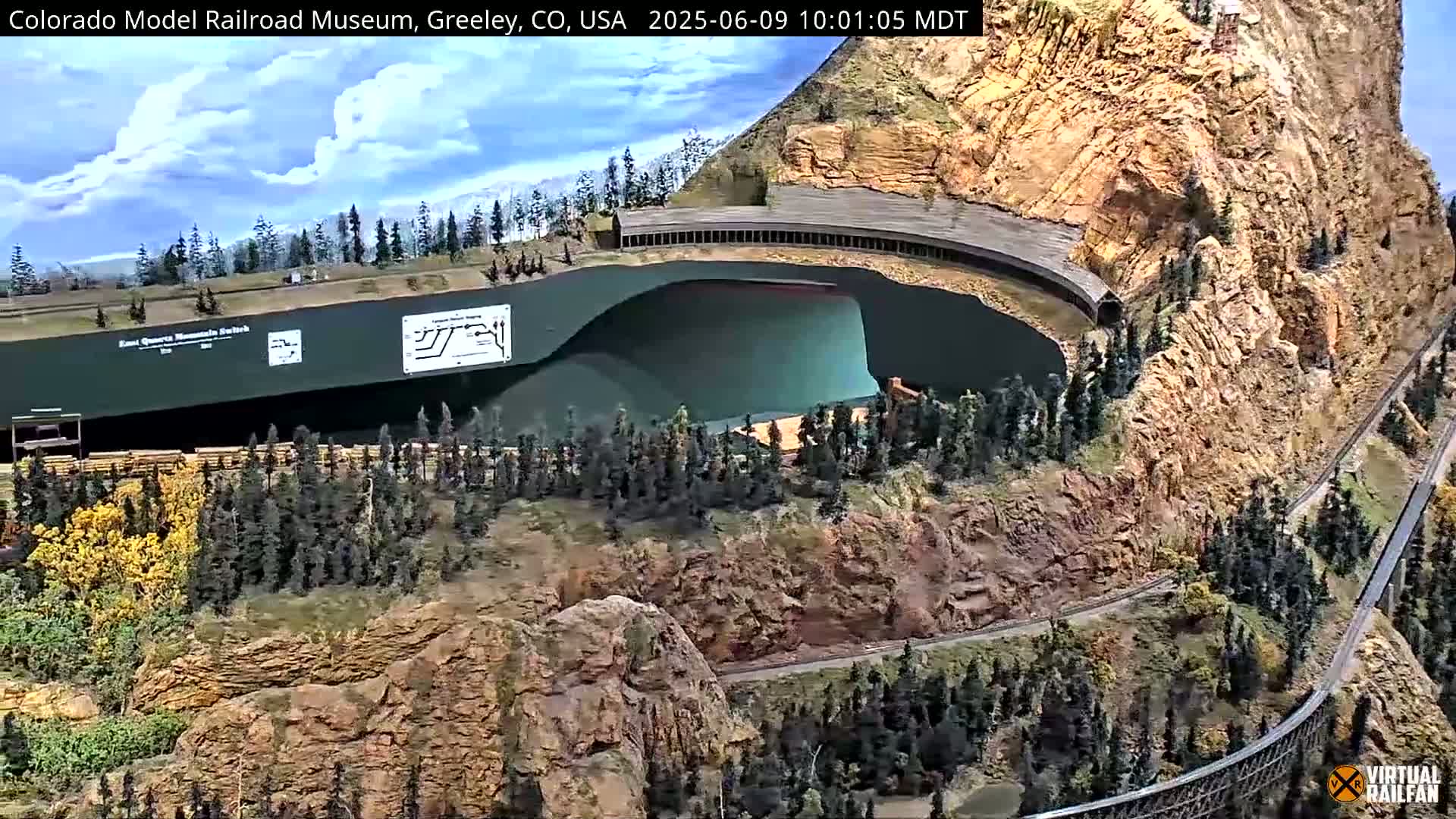 A large model train set depicting a mountainous landscape with a winding track, a lake, and a large structure resembling a tunnel or building carved into a cliffside, under partly cloudy skies.