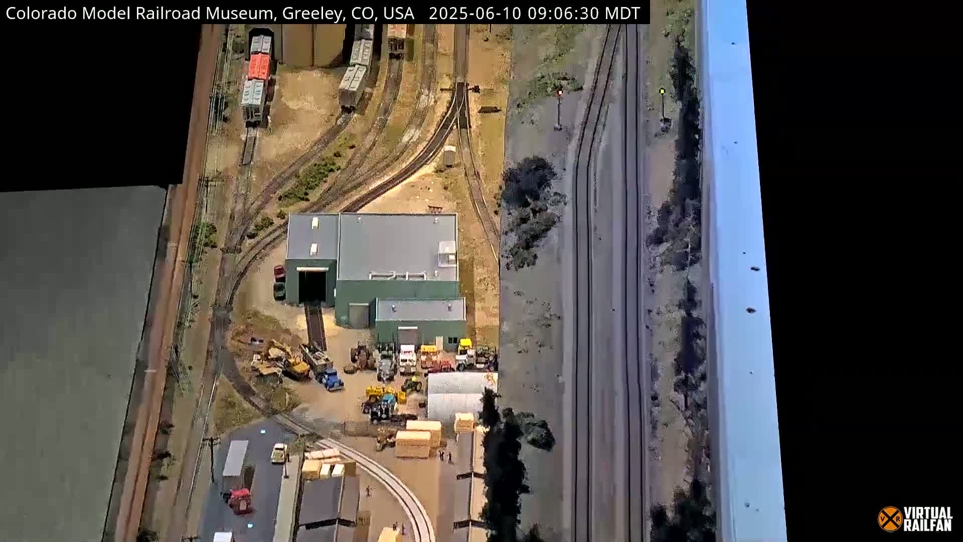 An aerial view shows a model train setup with various buildings, tracks, and vehicles, possibly under sunny conditions.