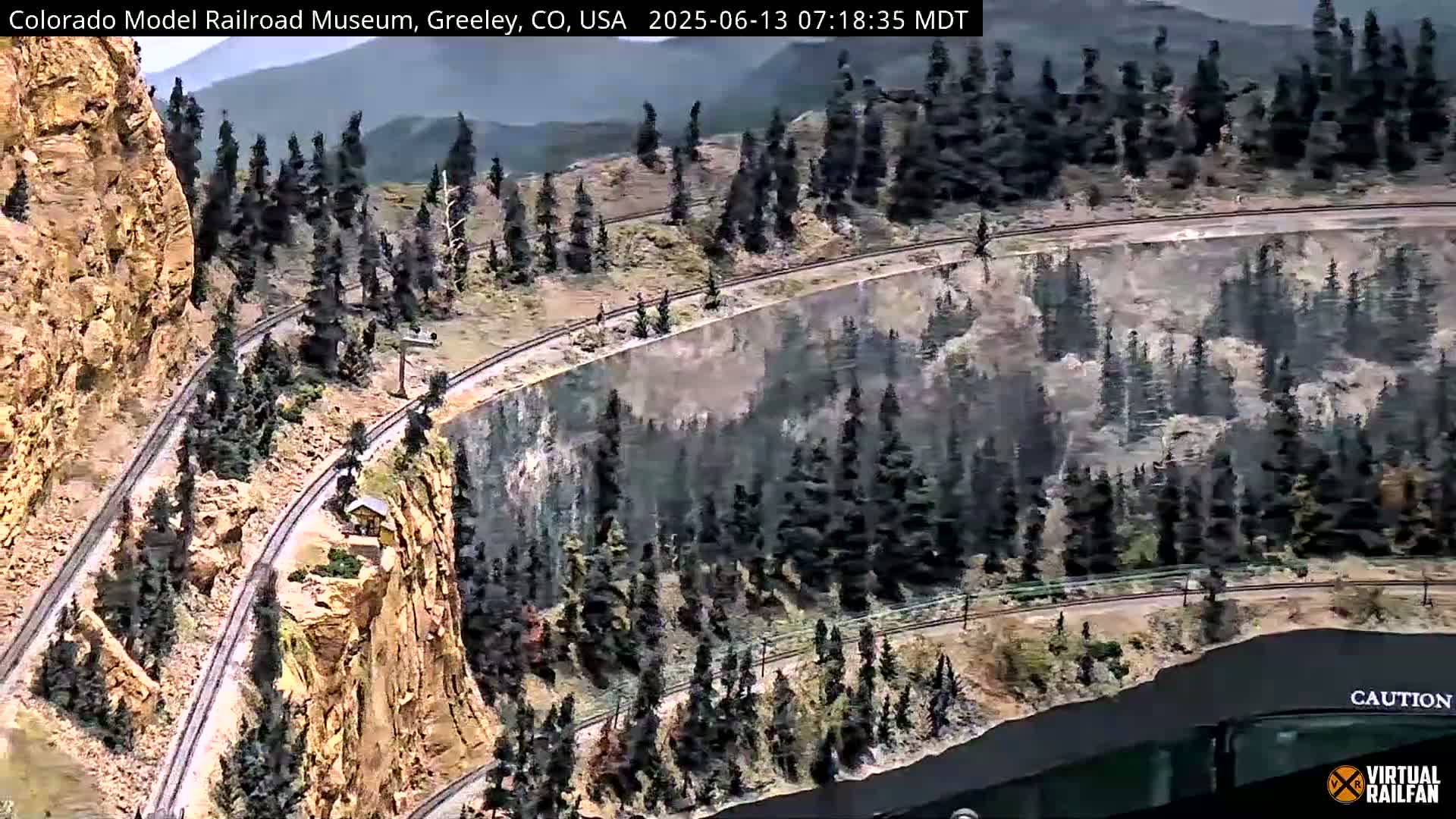 An overhead view of a detailed model railroad winding through a mountainous landscape with evergreen trees.