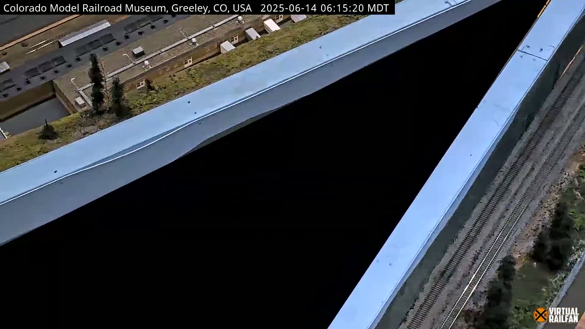 An overhead view shows a dark, V-shaped area between two light-blue structures, with model train tracks and landscaping visible below.