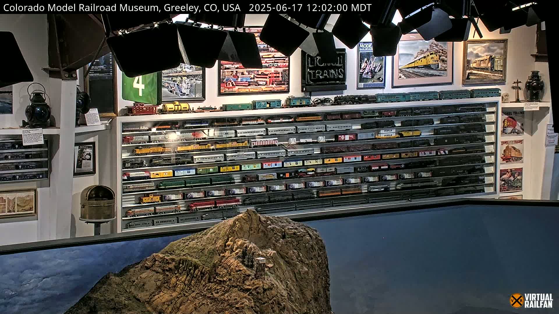 A model railroad museum display showcases numerous model trains in glass cases, with framed artwork and other railroad memorabilia on the walls, all visible behind a large model of a rocky mountain.