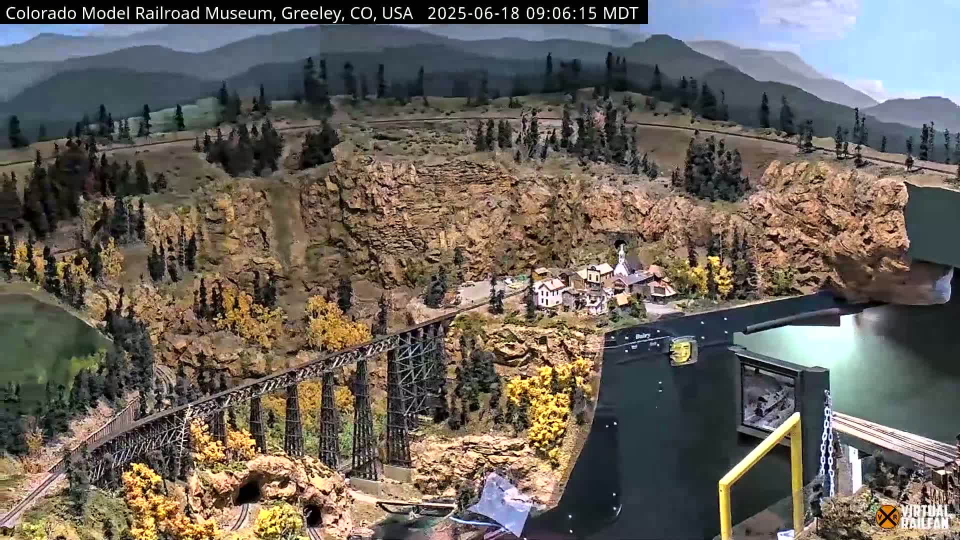A detailed model railroad, set in a mountainous landscape under a sunny sky, features a large wooden trestle bridge, a small town, and a rocky gorge.